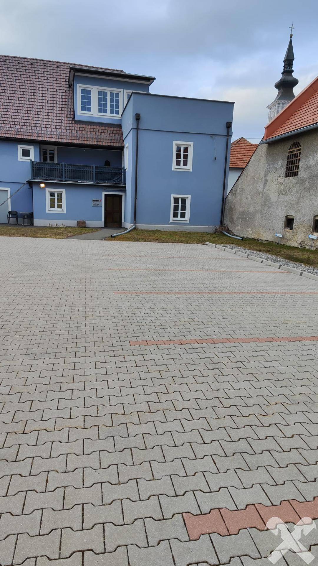 Gepflasterter Parkplatz vor dem blauen Gebäude mit einem Kirchturm in der Ferne.