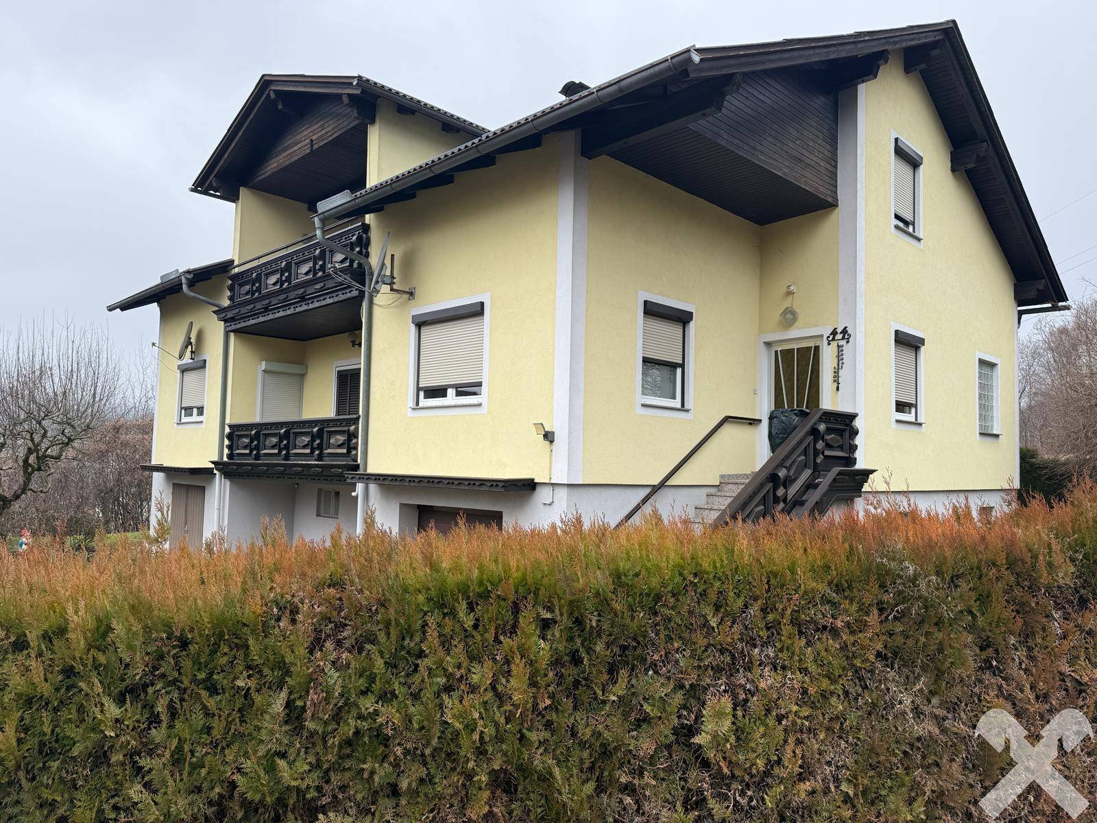 Ansicht des gelben Hauses von der Seite mit Außentreppe und Hecke im Vordergrund.