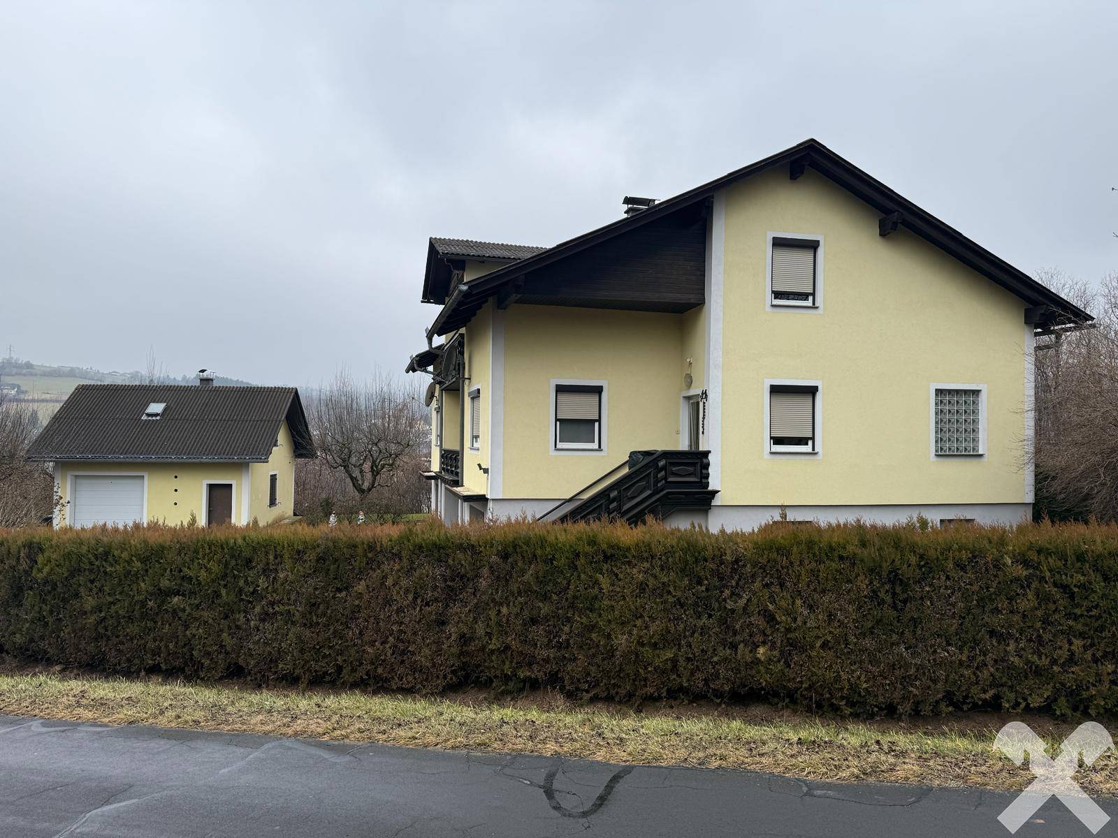 Seitenansicht des gelben Hauses mit Nebengebäude und einer dichten Hecke.