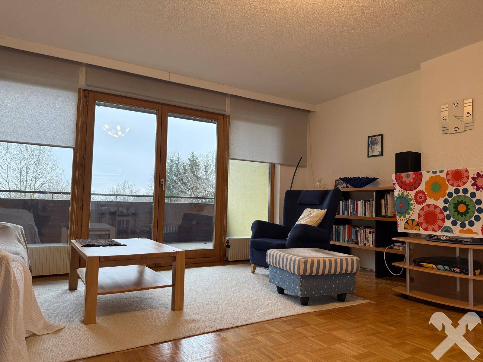 Wohnzimmer mit Parkettboden, großem Fenster und Zugang zum Balkon.