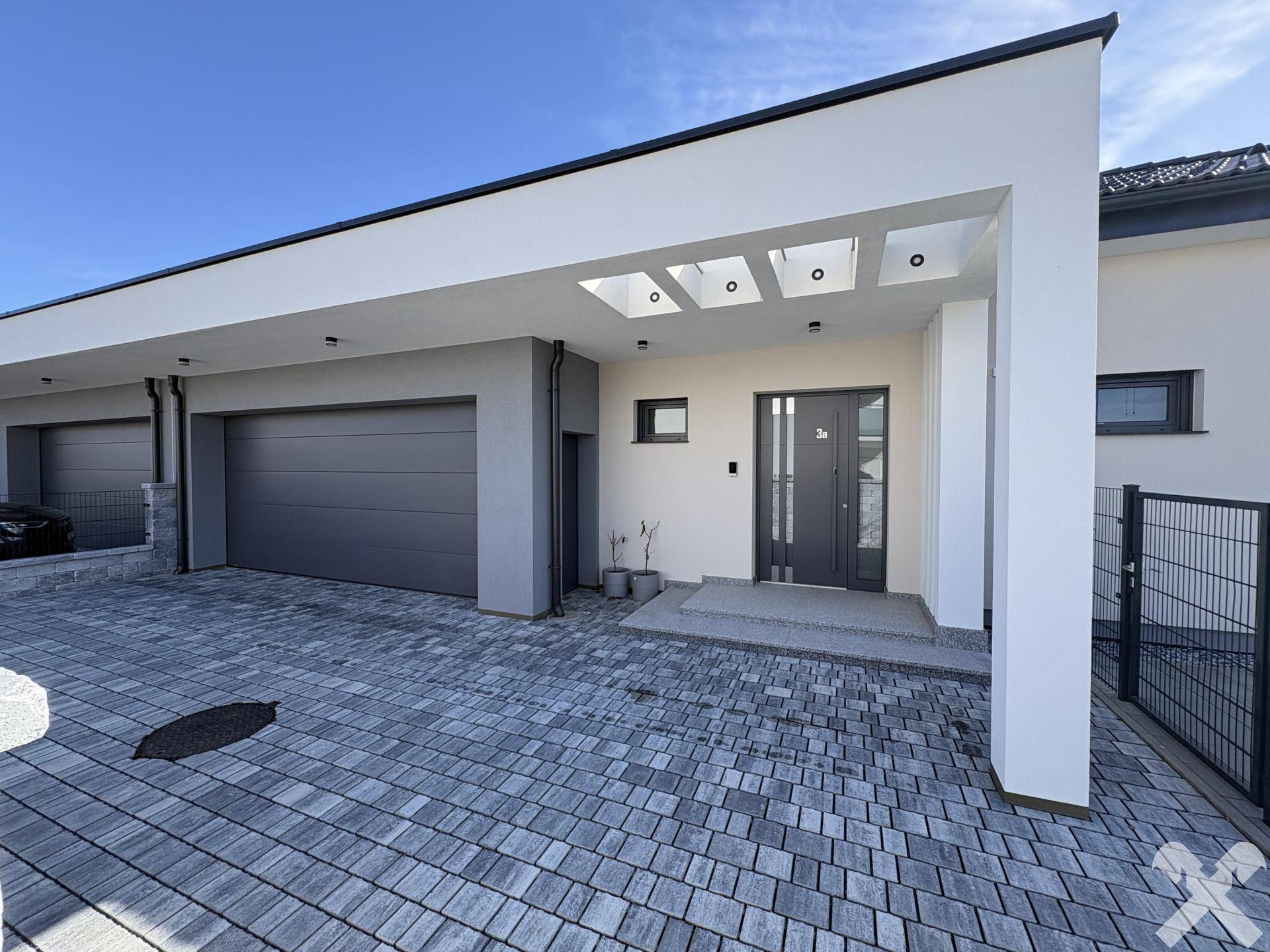 Moderne Hausfront mit integrierter Garage, stilvollem Eingang und gepflasterter Zufahrt bei Sonnenschein.