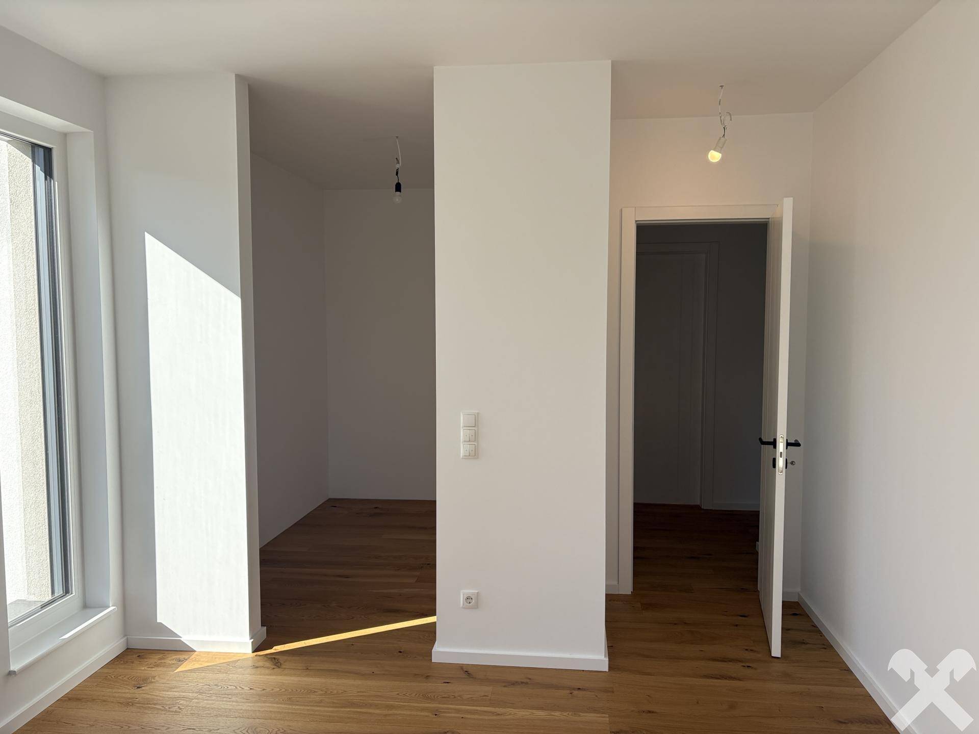 Helles Zimmer mit Holzboden und offener Tür, die zu einem weiteren Raum führt.