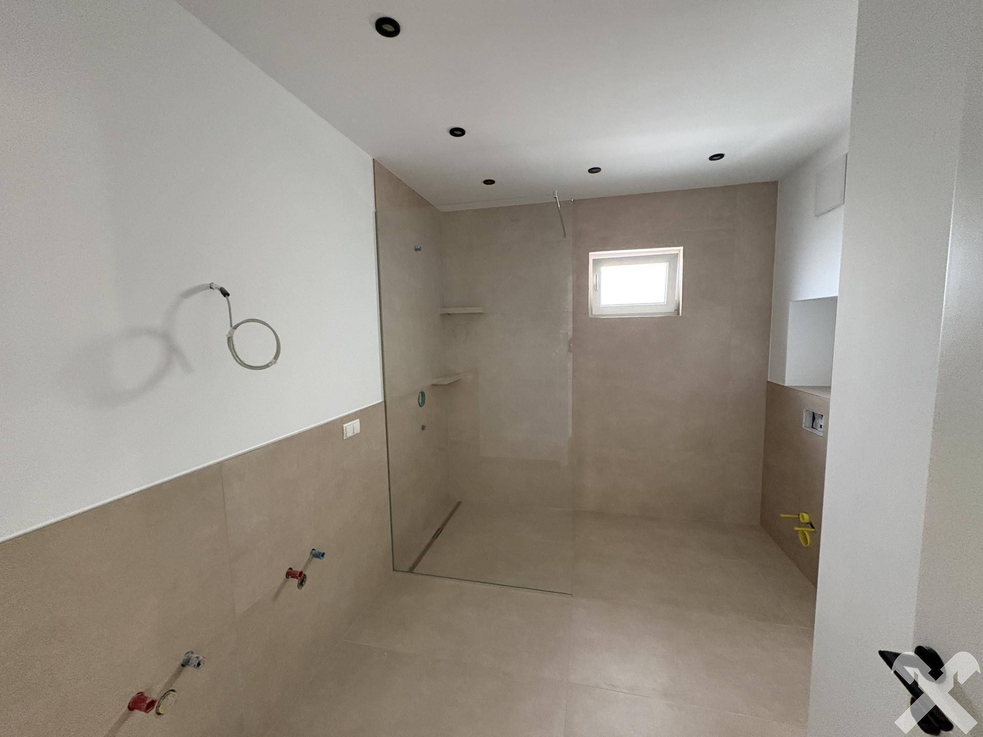 Geräumiges Badezimmer im Rohbau mit vorbereiteten Anschlüssen und einem Fenster für Tageslicht.