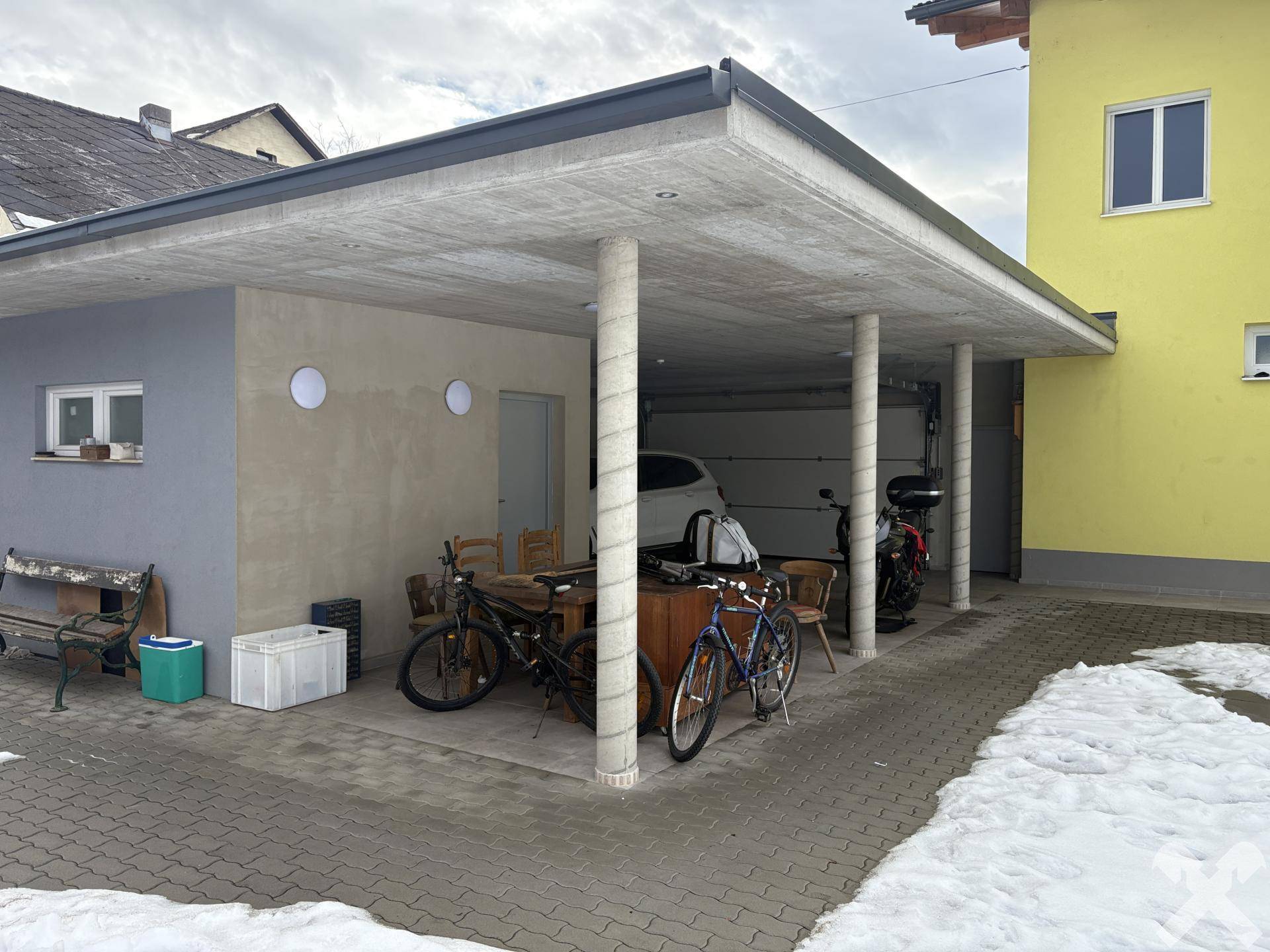 Überdachter Stellplatz mit Säulen, der auch als Abstellfläche für Fahrräder und Möbel dient.