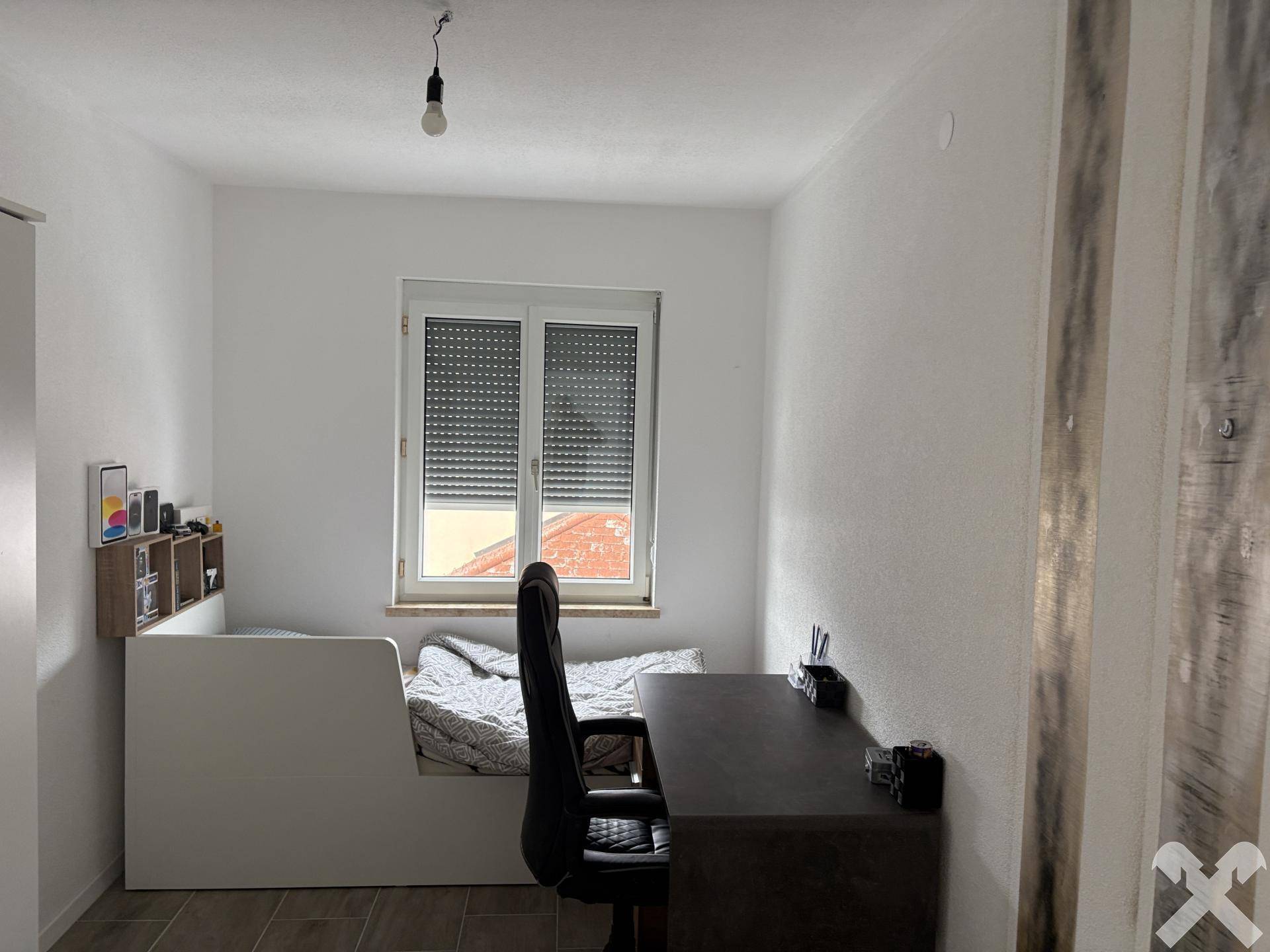 Helles Schlafzimmer mit Einzelbett, Schreibtisch und Fenster mit Rollladen.
