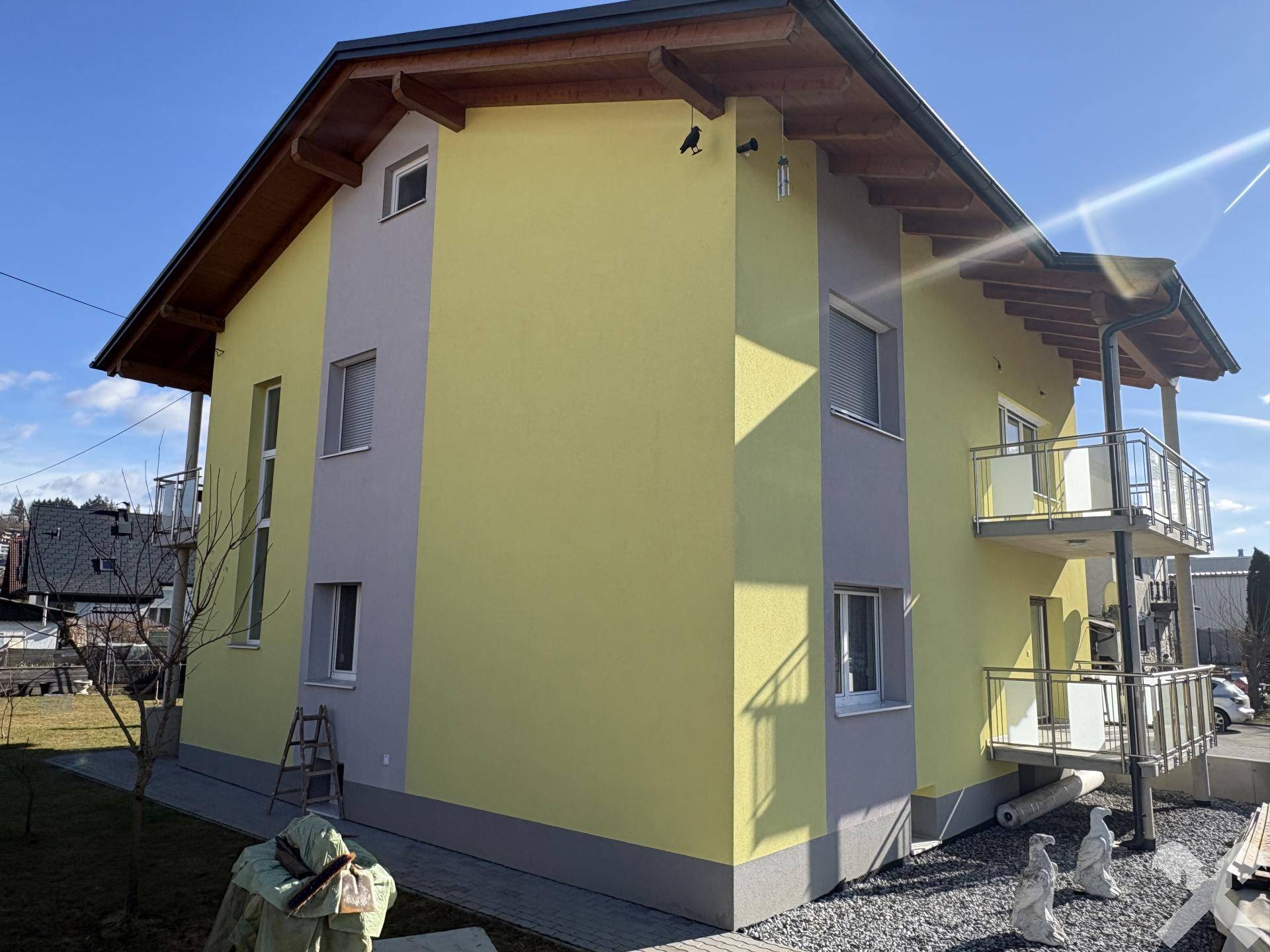 Seitenansicht des gelben Hauses mit Balkonen und einem kleinen Gartenbereich bei Sonnenschein.