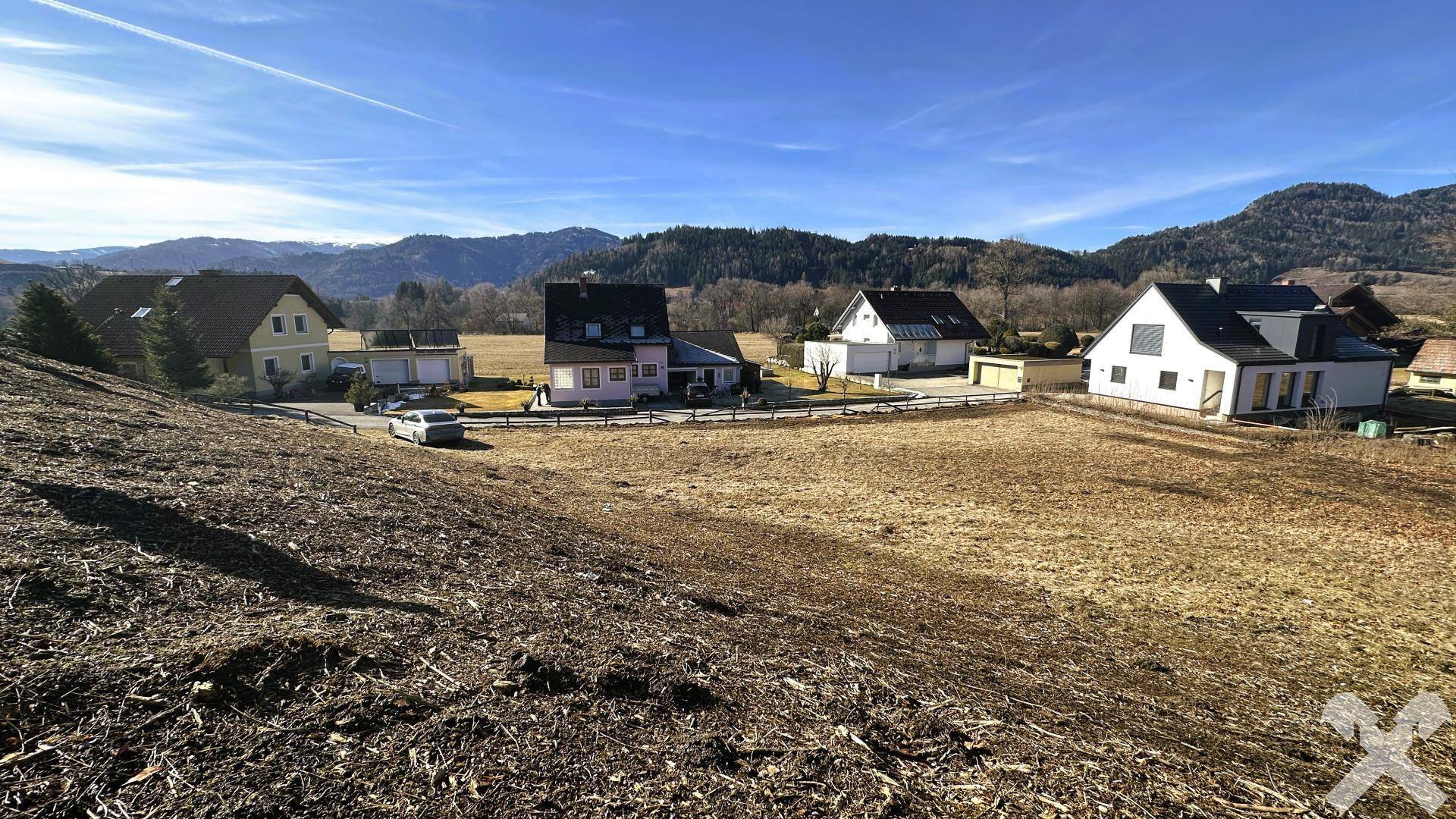 Offenes Grundstück mit mehreren Wohnhäusern im Hintergrund und einer weiten Bergkulisse.