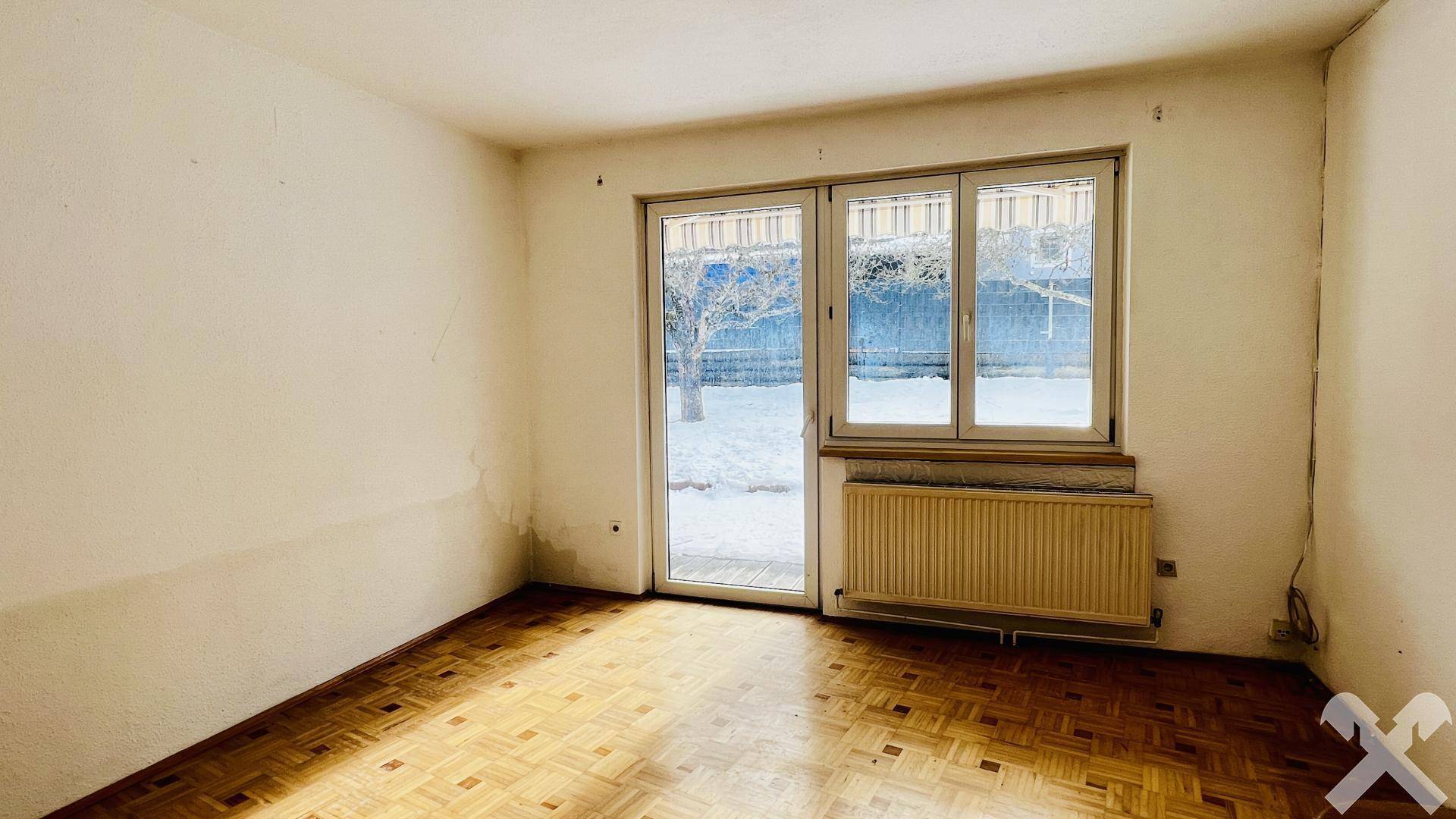 Wohnzimmer mit Parkettboden, Heizkörper und Zugang zum Garten durch große Terrassentür.
