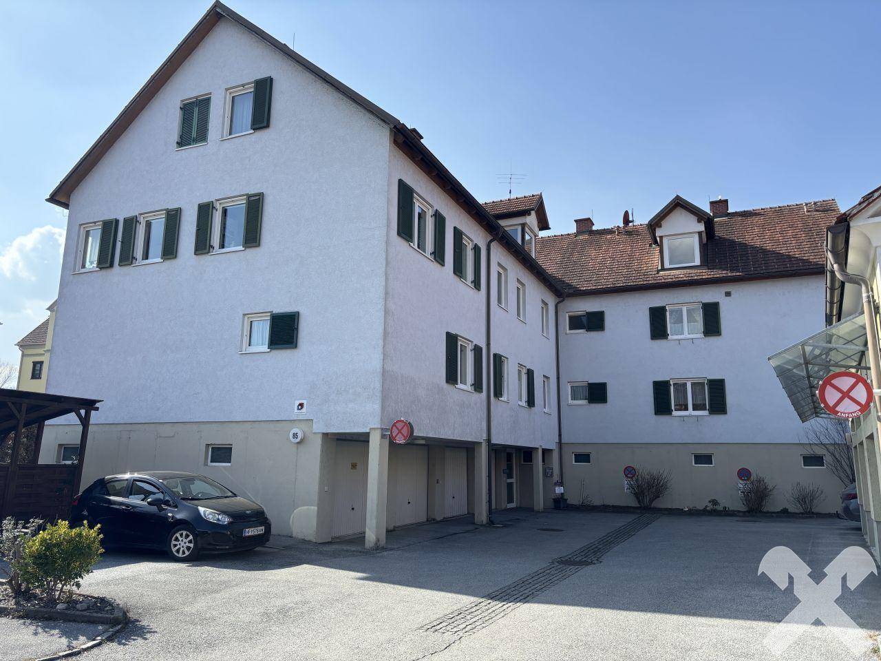 Mehrfamilienhaus mit Parkplätzen und Garagen im Erdgeschoss unter blauem Himmel.