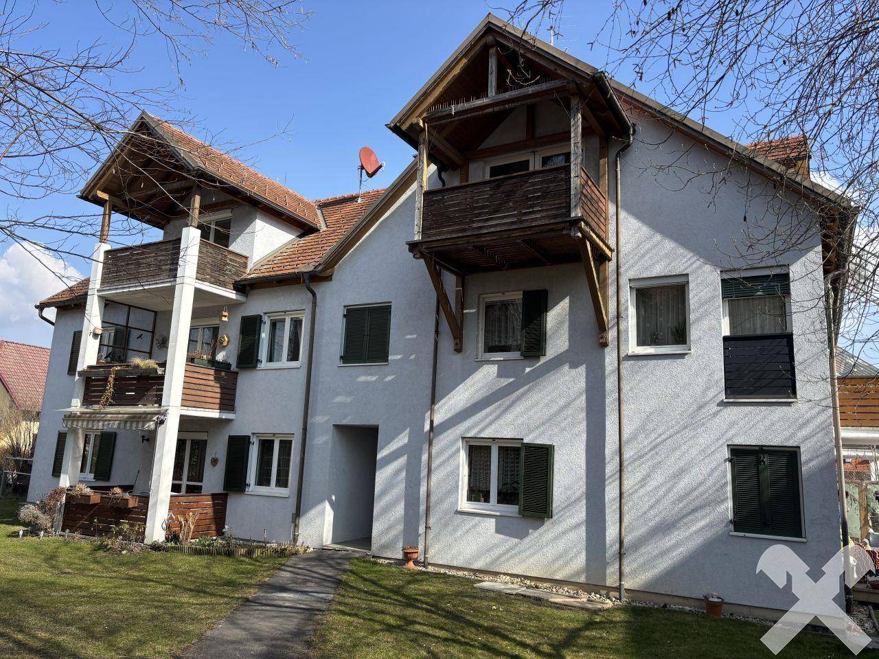 Mehrfamilienhaus mit Balkonen, gepflegtem Vorgarten und traditioneller Architektur unter blauem Himmel.