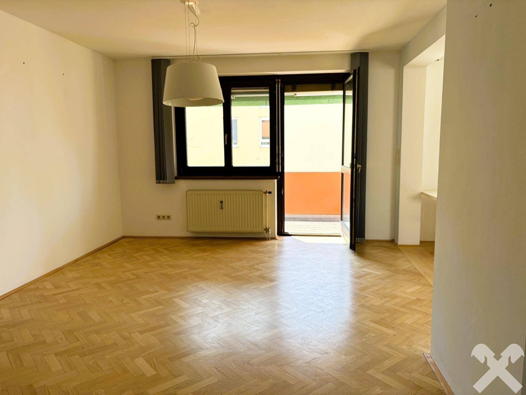 Geräumiger Wohnbereich mit hellem Parkettboden und großem Fenster, das viel Sonnenlicht hereinlässt.