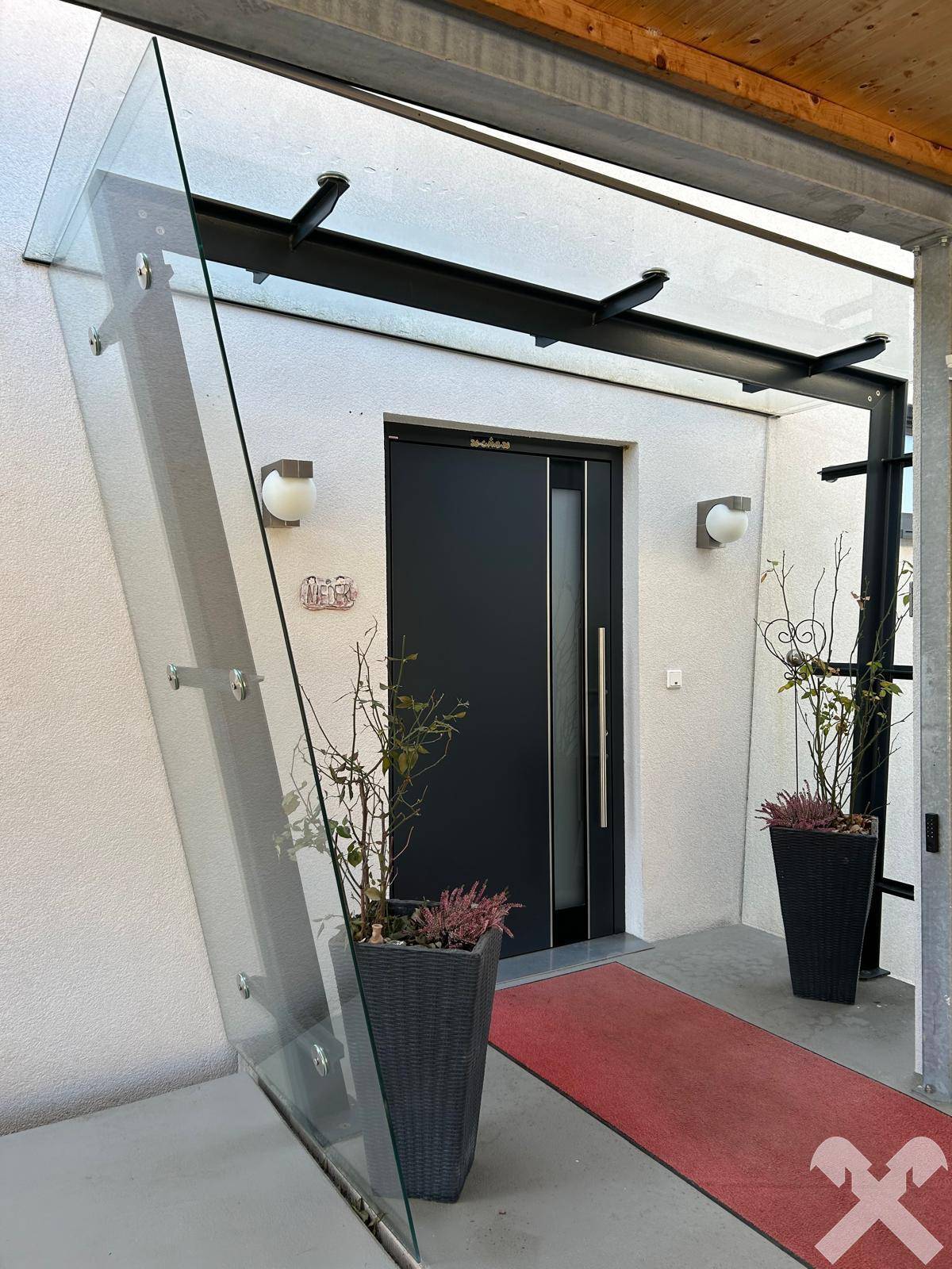 Moderner Eingangsbereich mit Glasvordach und dunkler Haustür, umgeben von schlichten Pflanzen.