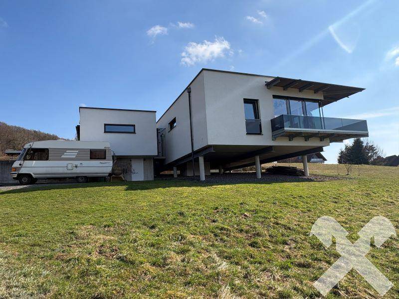 Architektonisch interessantes Haus mit großen Fensterfronten und Wohnmobil-Stellplatz auf grünem Hügel.
