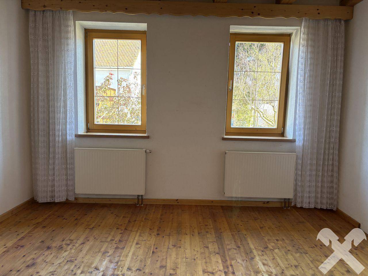 Leerer Raum mit Holzboden, zwei Fenstern und Heizkörpern unter der Holzbalkendecke.