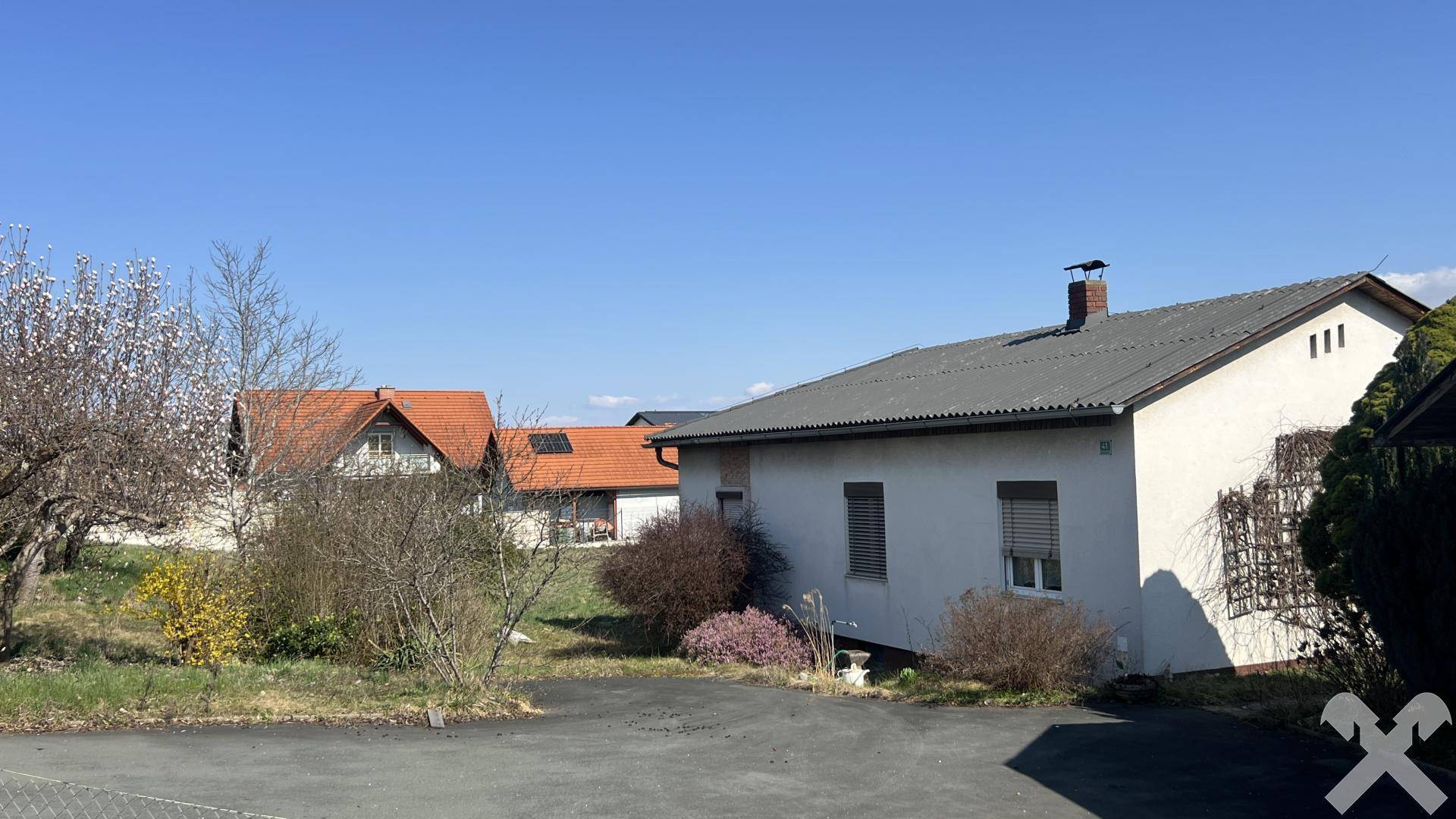 Einfamilienhaus mit weißer Fassade und grauen Dachziegeln, umgeben von Sträuchern und Bäumen.