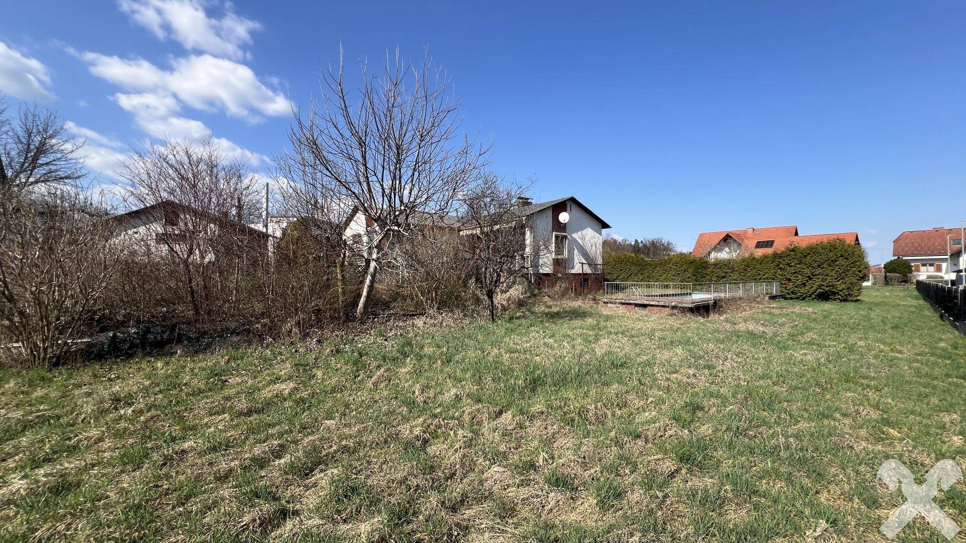 Offenes Grundstück mit Gras und Bäumen, im Hintergrund ein Haus mit Holzterrasse und Hecke.