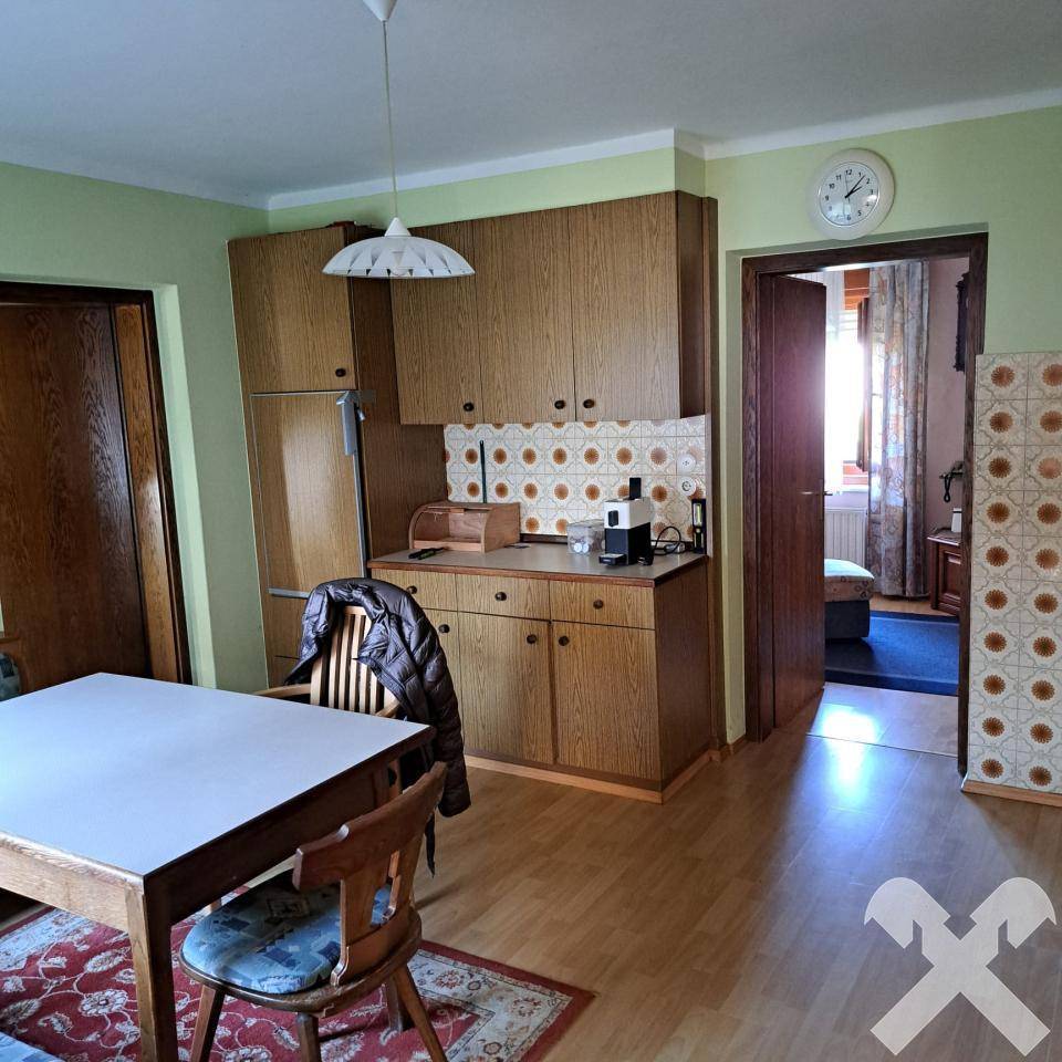 Einfache Küche mit Holzschränken, Esstisch und Blick in einen weiteren Raum.
