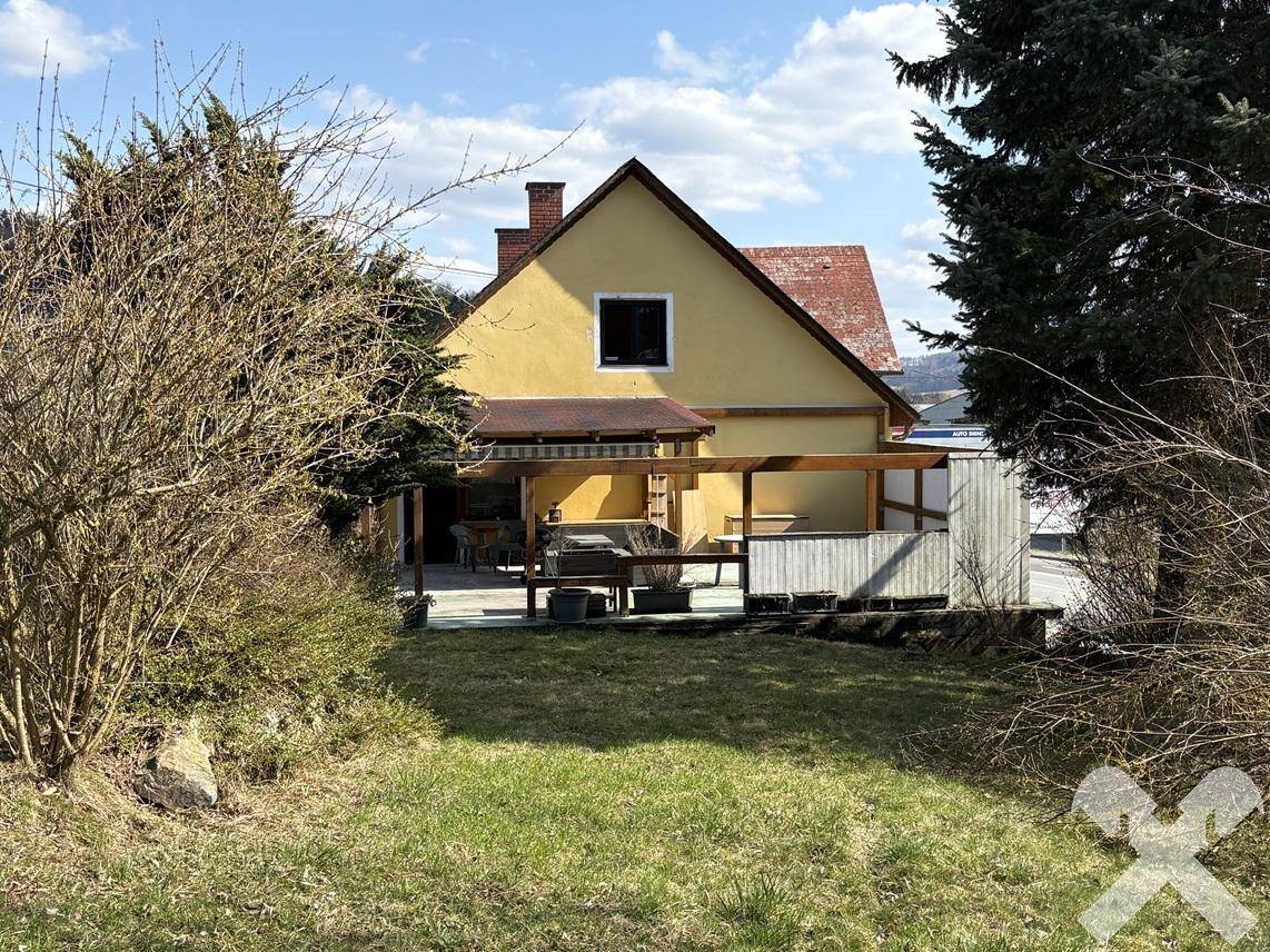 Gartenbereich hinter dem Haus mit Rasen, Bäumen und einer überdachten Terrasse mit Sitzgelegenheiten.