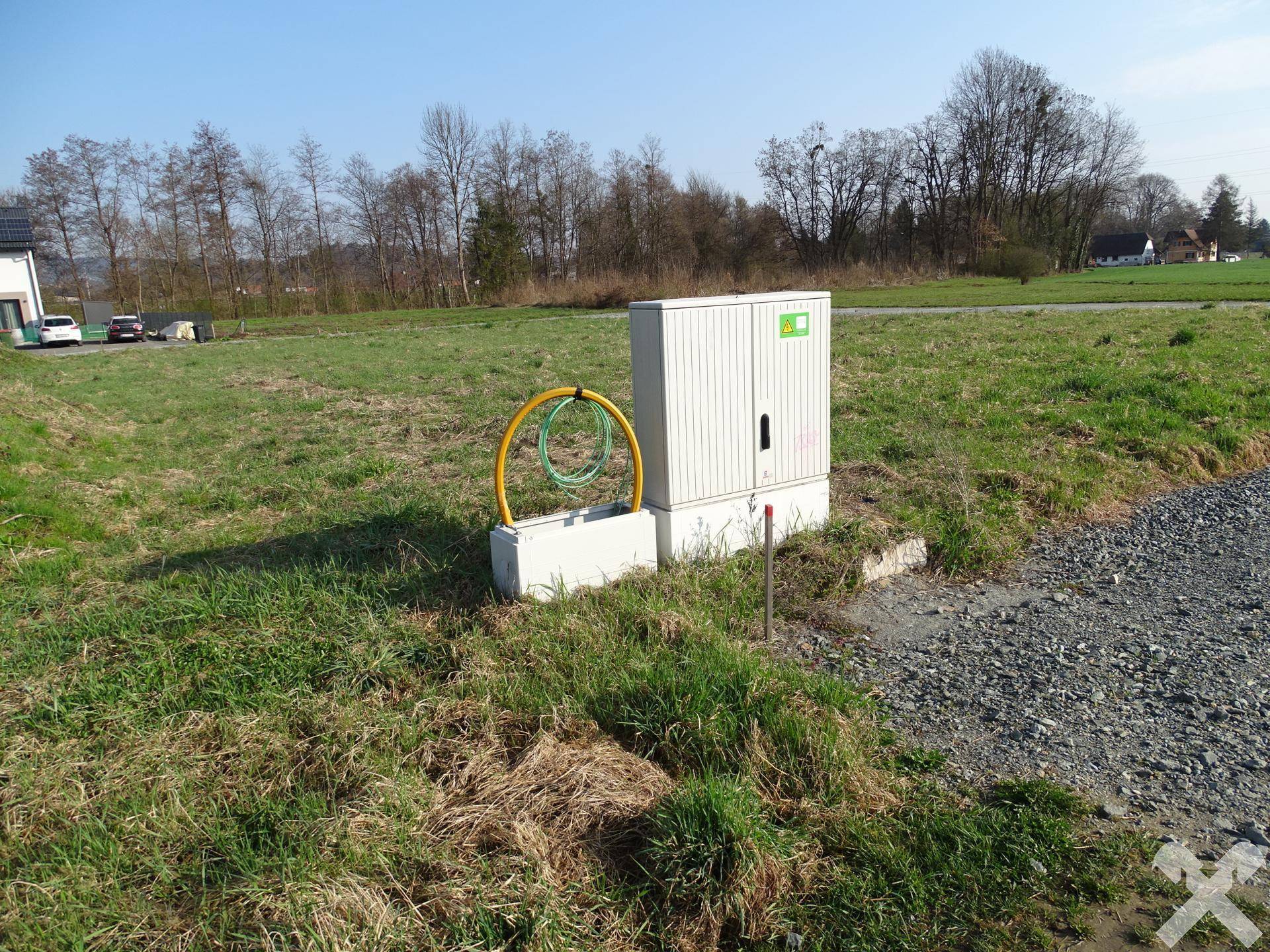 Versorgungskasten mit gelben Rohren auf Betonfundament neben Schotterweg und Grasfläche.