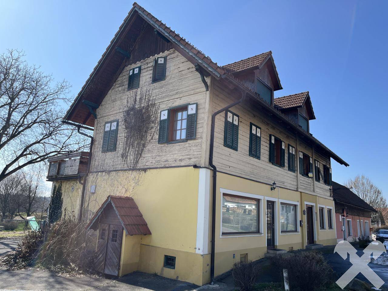 Traditionelles Gebäude mit gelber Fassade, Holzverkleidung und grünen Fensterläden unter blauem Himmel.