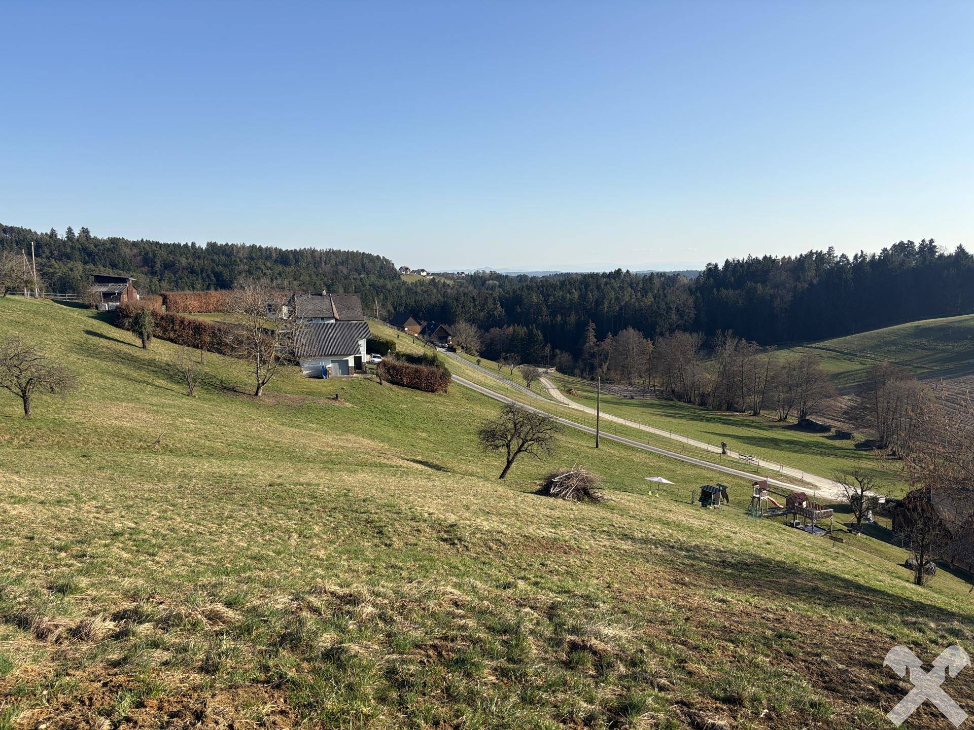 Weitläufige Hügellandschaft mit vereinzelten Häusern und Wald im Hintergrund.