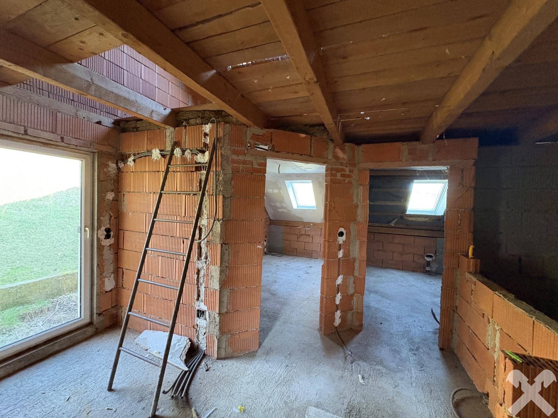 Dachgeschoss im Rohbau mit Ziegelwänden, Holzbalken und großen Fenstern.