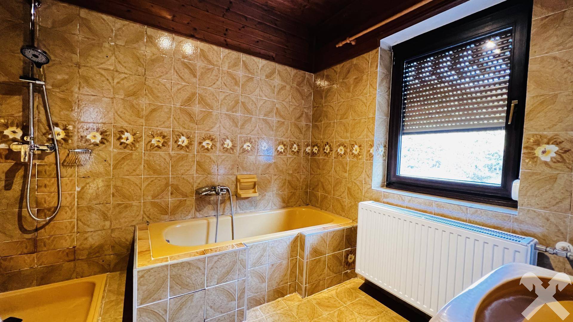 Badezimmer mit Badewanne, Dusche und Fenster, ausgestattet mit älteren Fliesen.