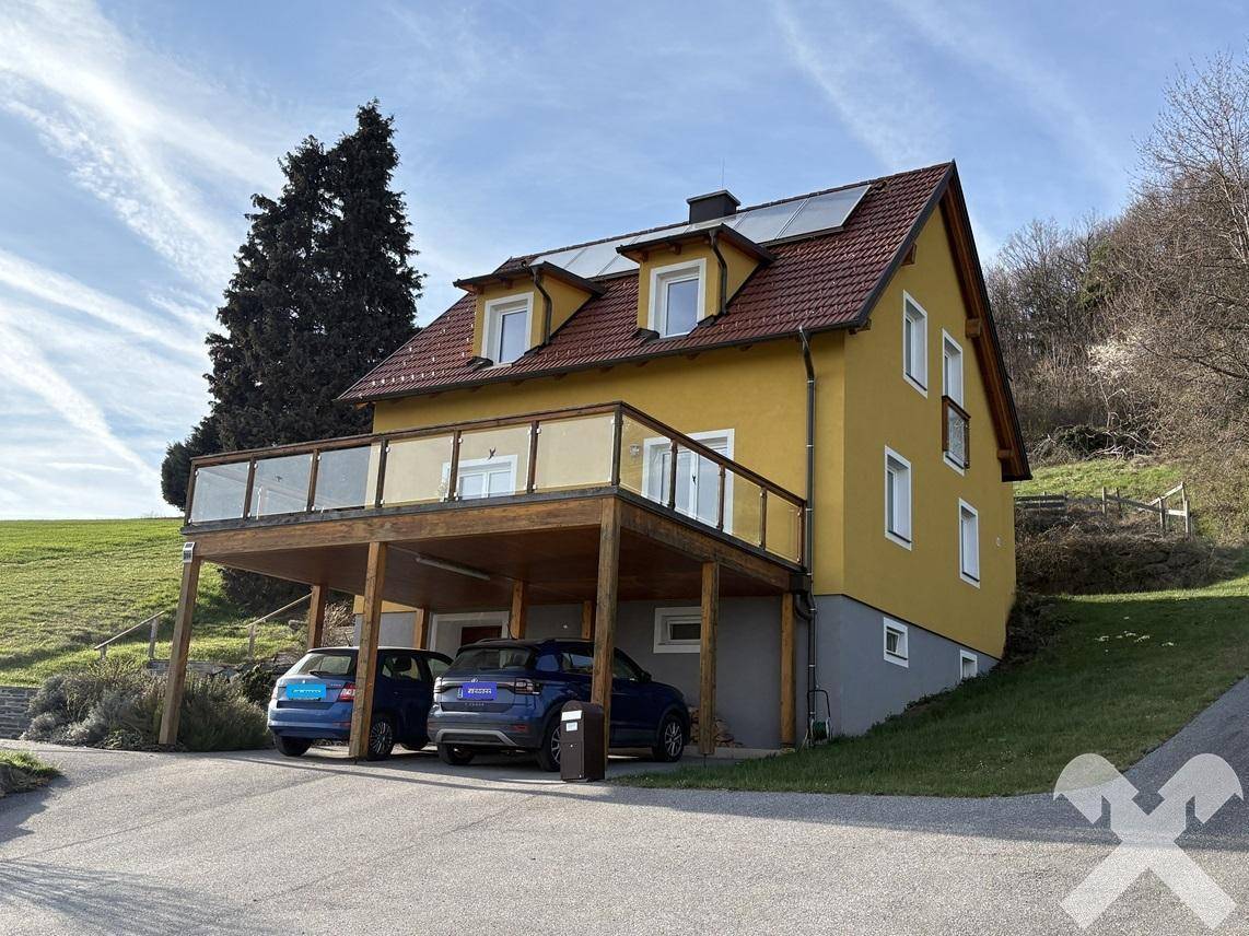 Ansicht des gelben Hauses mit Carport und Photovoltaikanlage, eingebettet in eine ländliche Umgebung.