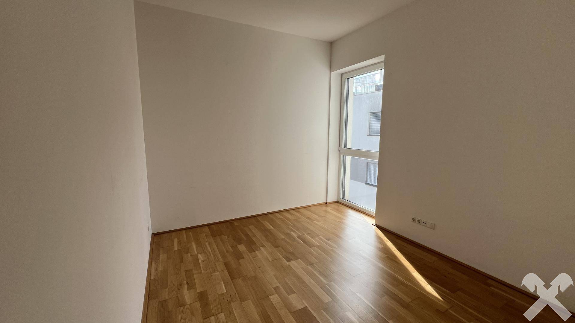 Helles, leeres Zimmer mit Holzparkett und einem hohen Fenster, das viel Sonnenlicht spendet.