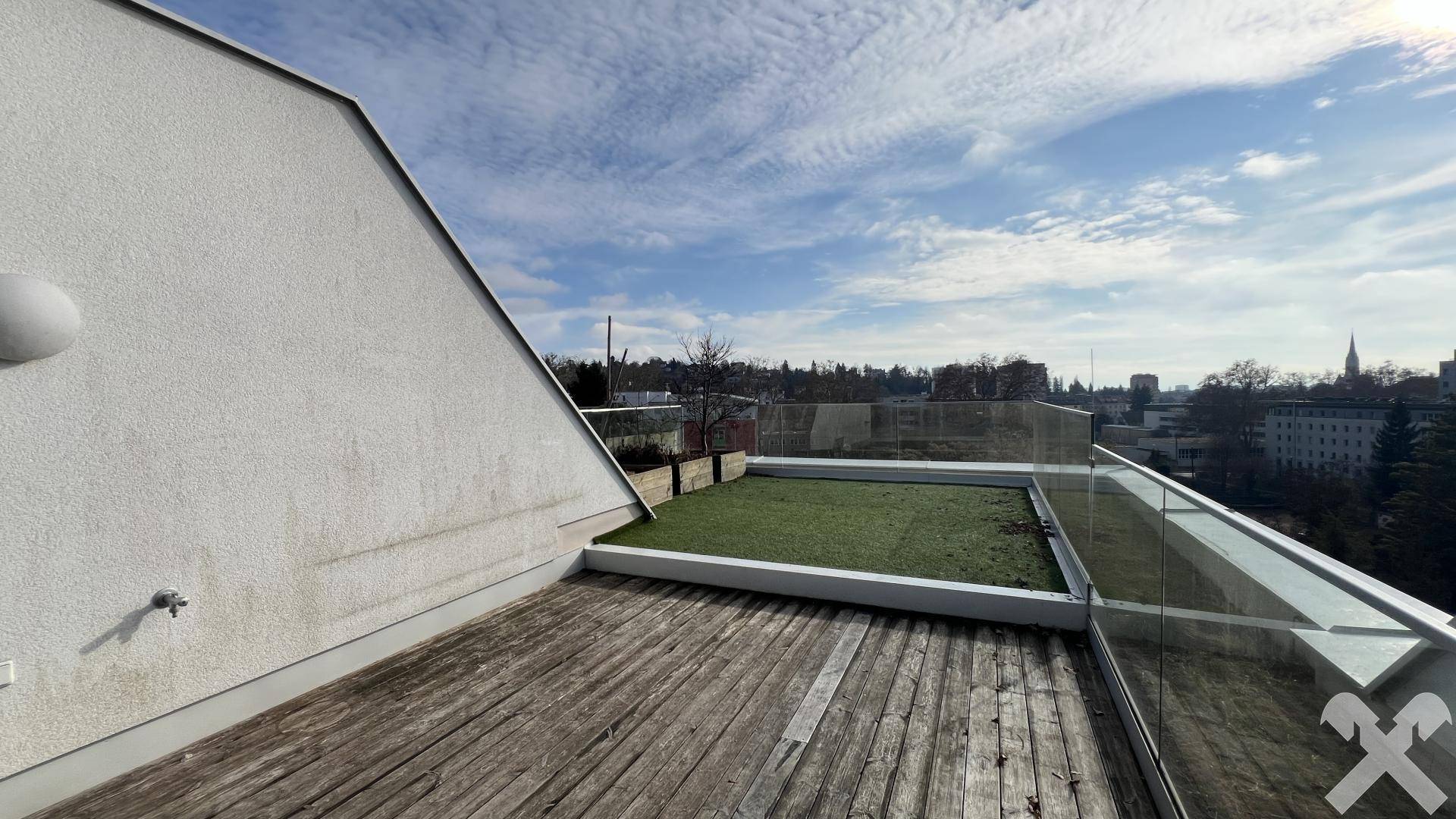 Weitläufige Dachterrasse mit Holzboden und Glasbrüstung unter blauem Himmel mit Wolken.