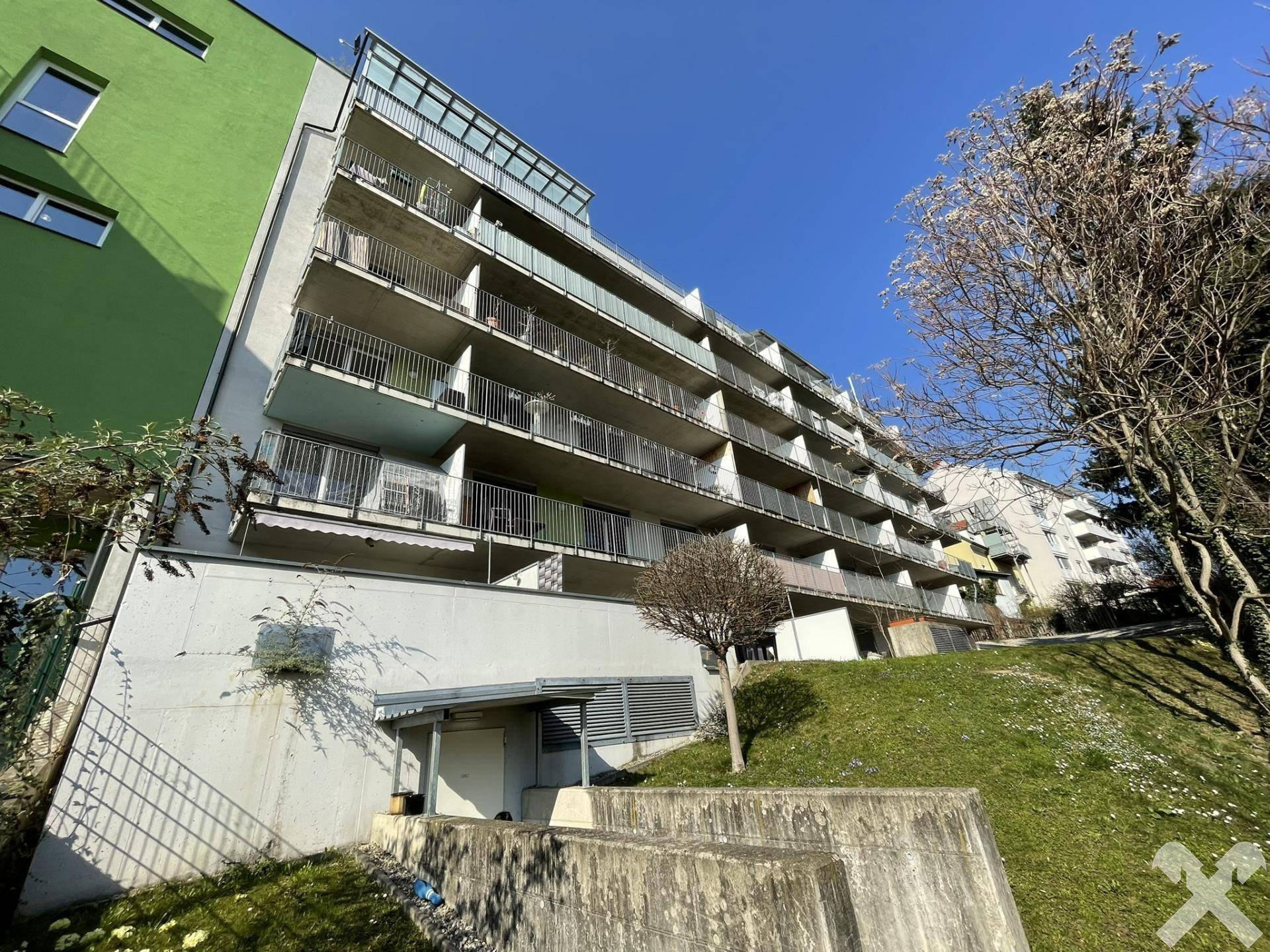 Mehrstöckiges Wohngebäude mit zahlreichen Balkonen und grüner Fassade an einem sonnigen Hang.