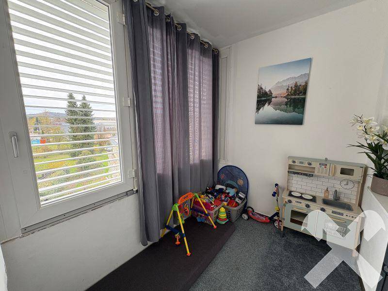 Helles Kinderzimmer mit Spielzeug, Teppichboden und großen Fenstern mit Ausblick ins Grüne.