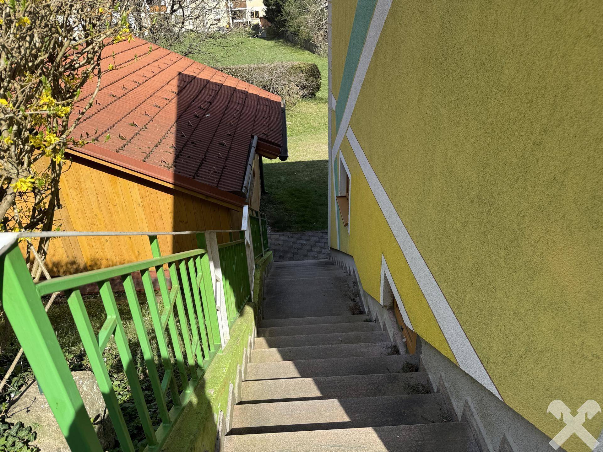 Außentreppe zwischen zwei Gebäuden mit grünem Geländer, führt zu einem oberen Bereich.