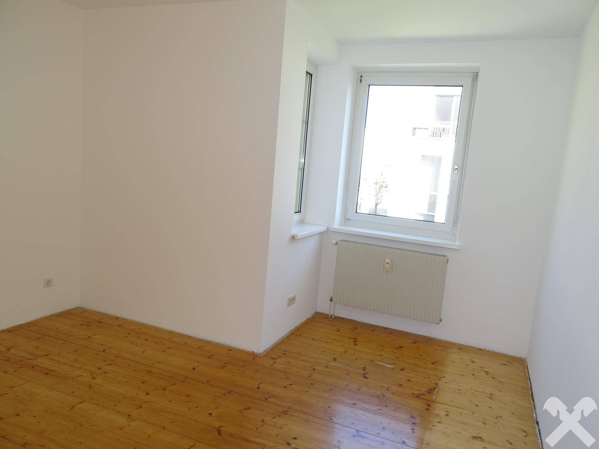 Leeres Zimmer mit Holzfußboden, weißen Wänden und großem Fenster für viel Licht.