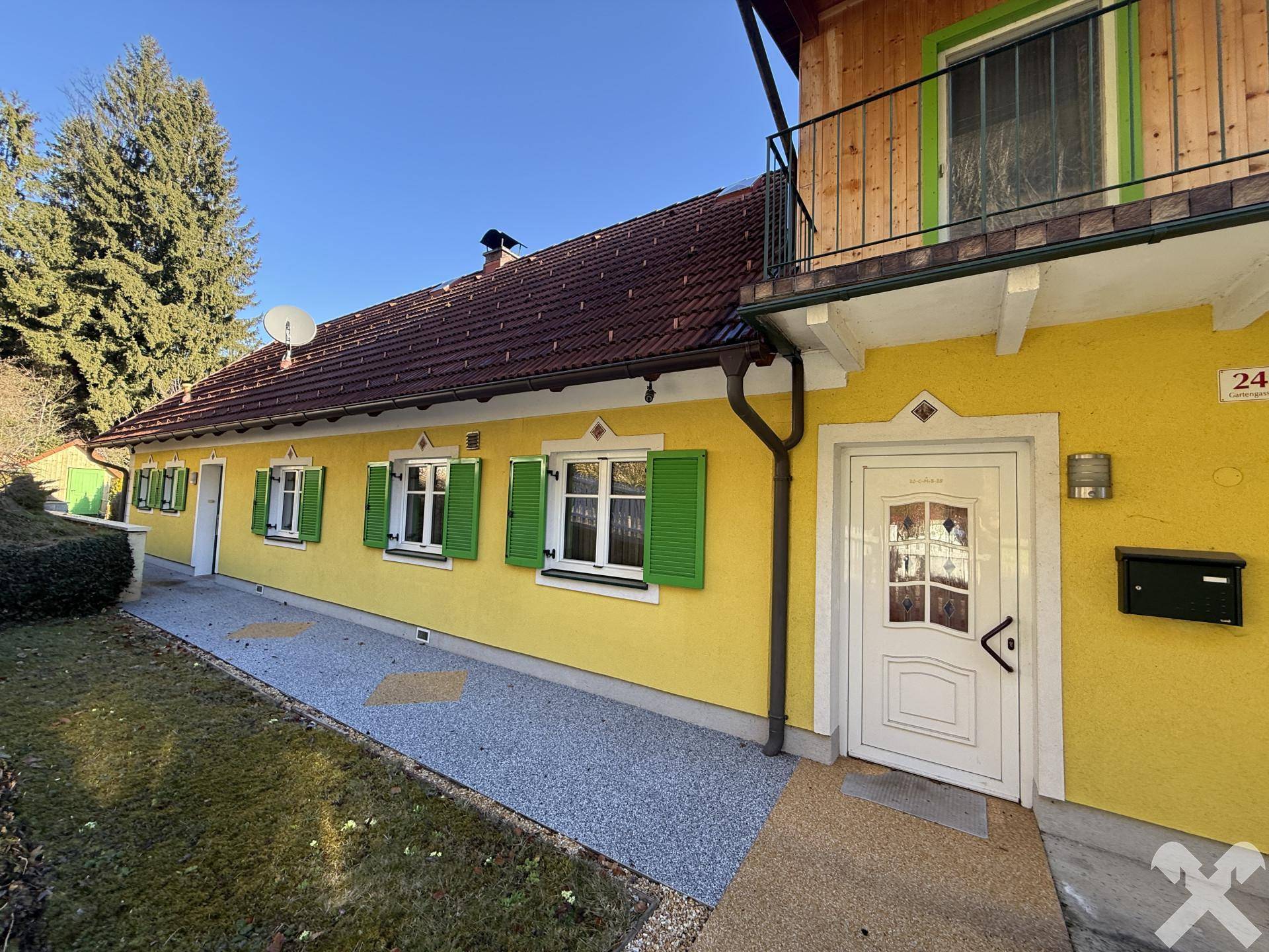 Gelbe Hausfassade mit grünen Fensterläden, weißer Eingangstür und gepflegtem Vorgarten.