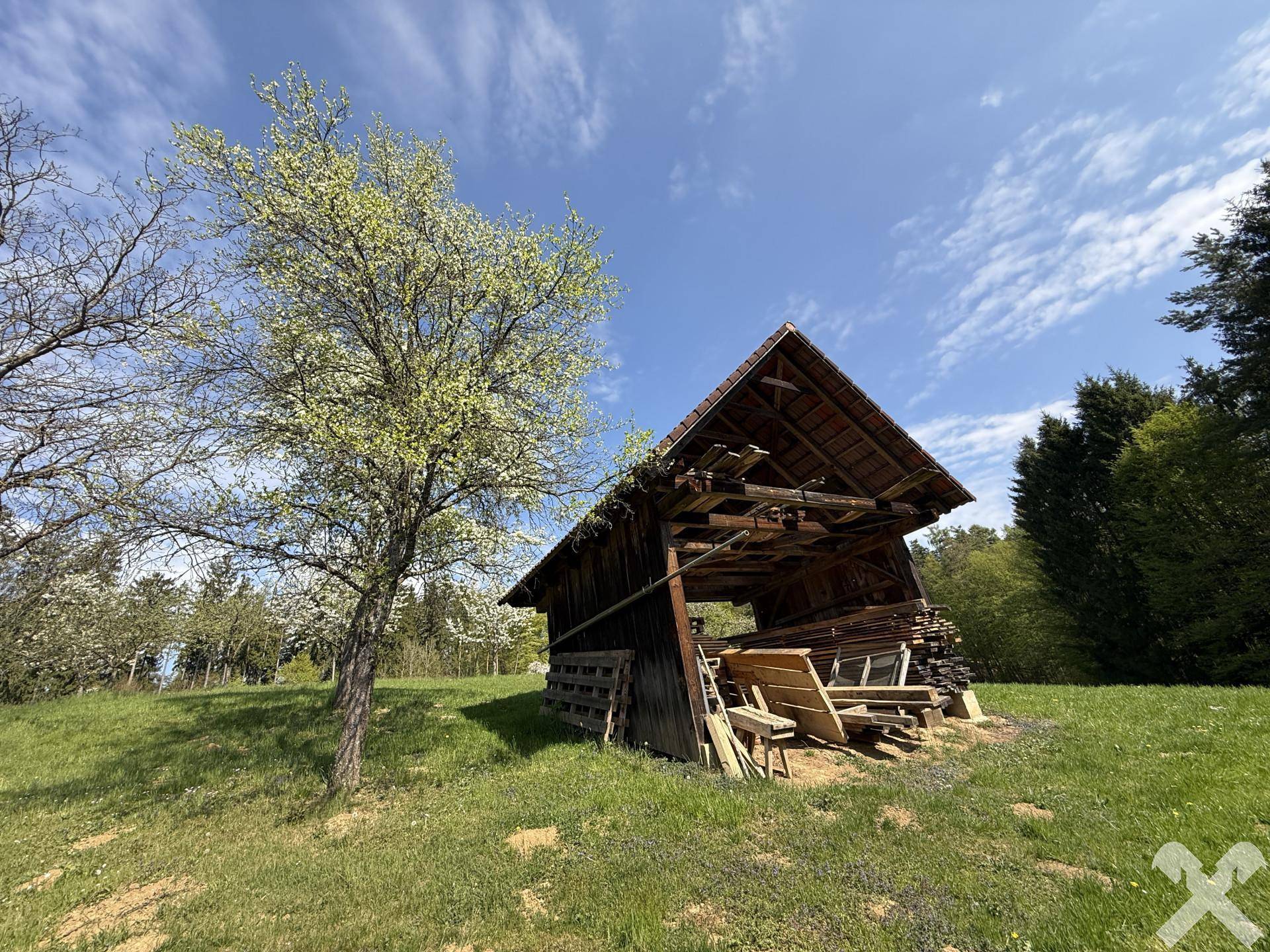 Rustikaler Holzschuppen mit offenem Dach und gelagertem Holz in einer grünen Wiese.