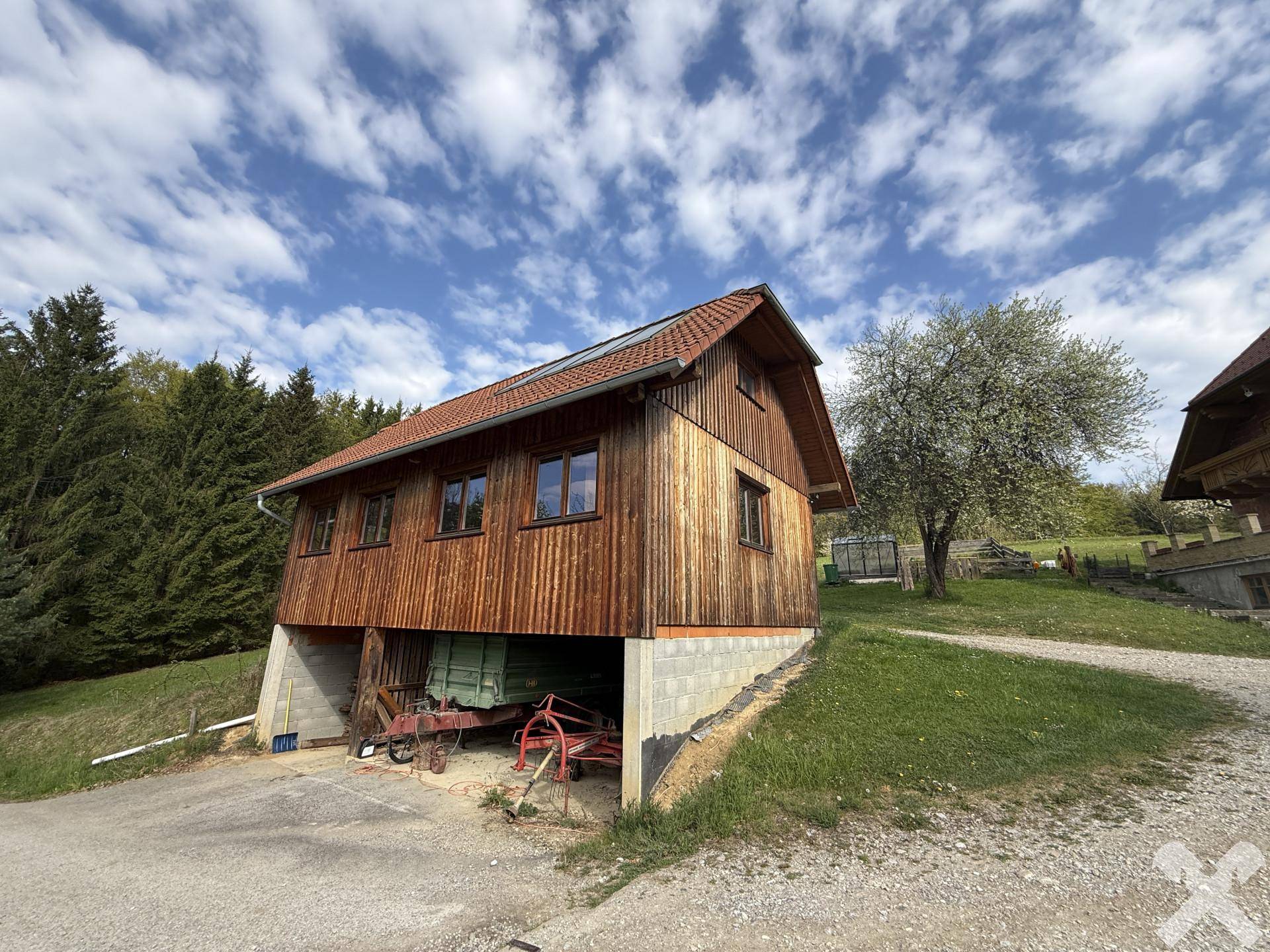 Holzgebäude mit Carport im Untergeschoss, umgeben von grüner Natur und Bäumen.