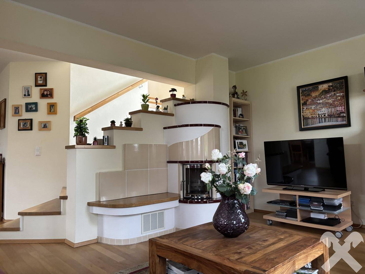 Wohnzimmer mit traditionellem Kachelofen, Holztreppe und Zugang zu weiteren Räumen.