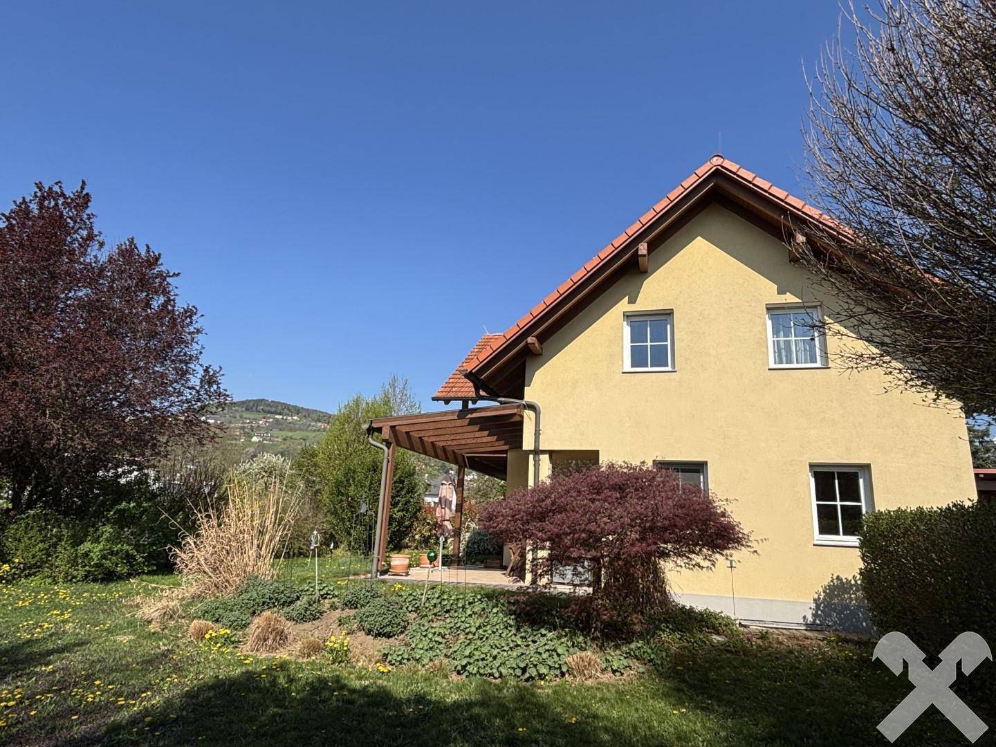 Seitlicher Blick auf das Haus mit überdachter Terrasse und blühendem Gartenbereich.