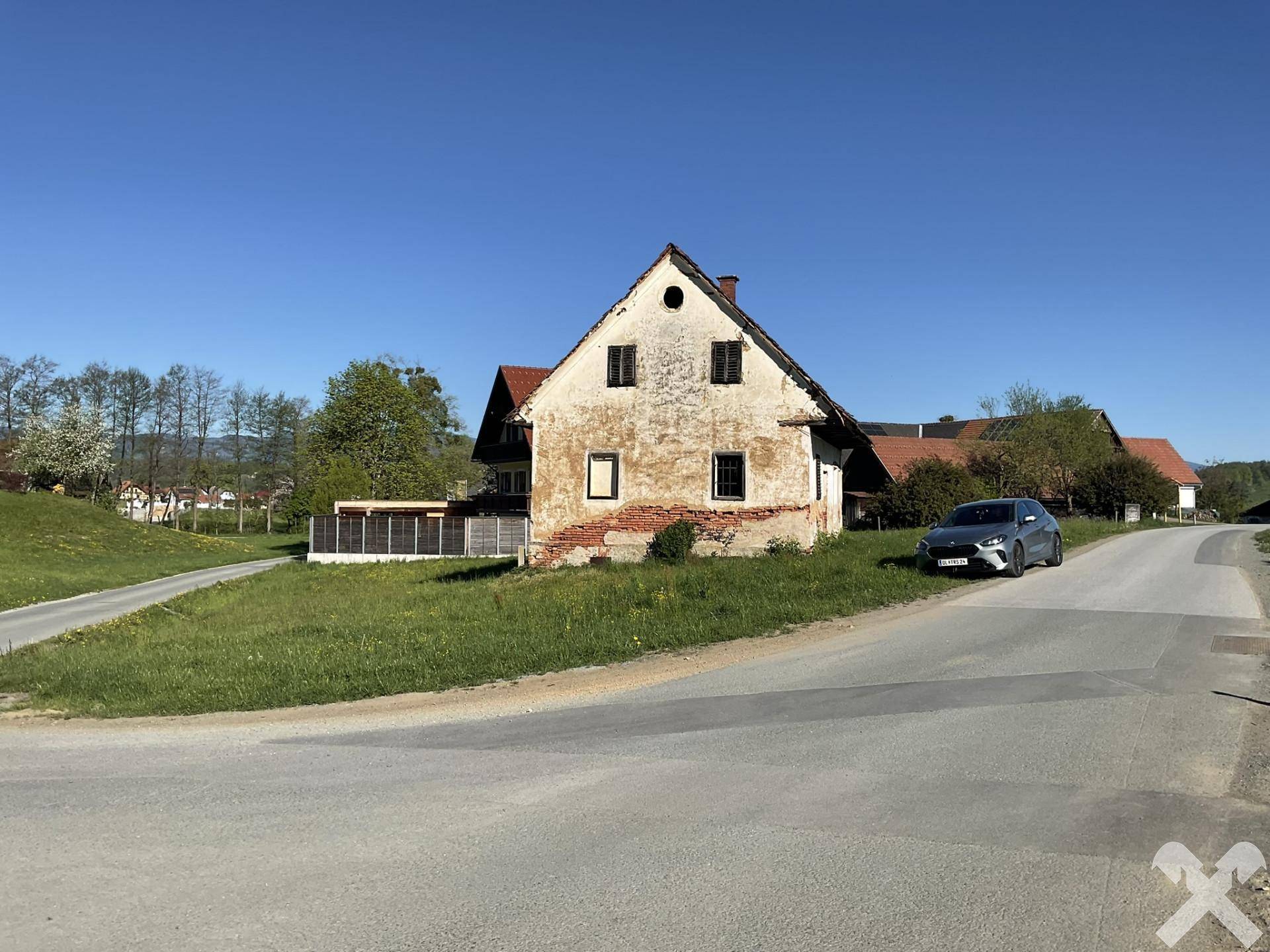 Gesamtansicht des alten Gebäudes in ländlicher Umgebung mit Zufahrtsstraße und weiteren Häusern im Hintergrund.