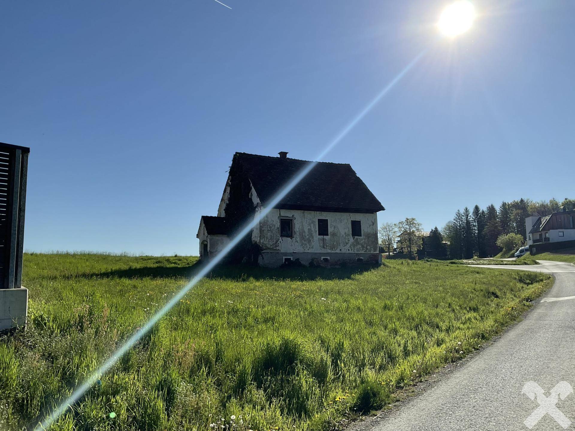 Altes Haus in sonniger Landschaft, umgeben von grünen Feldern unter blauem Himmel.