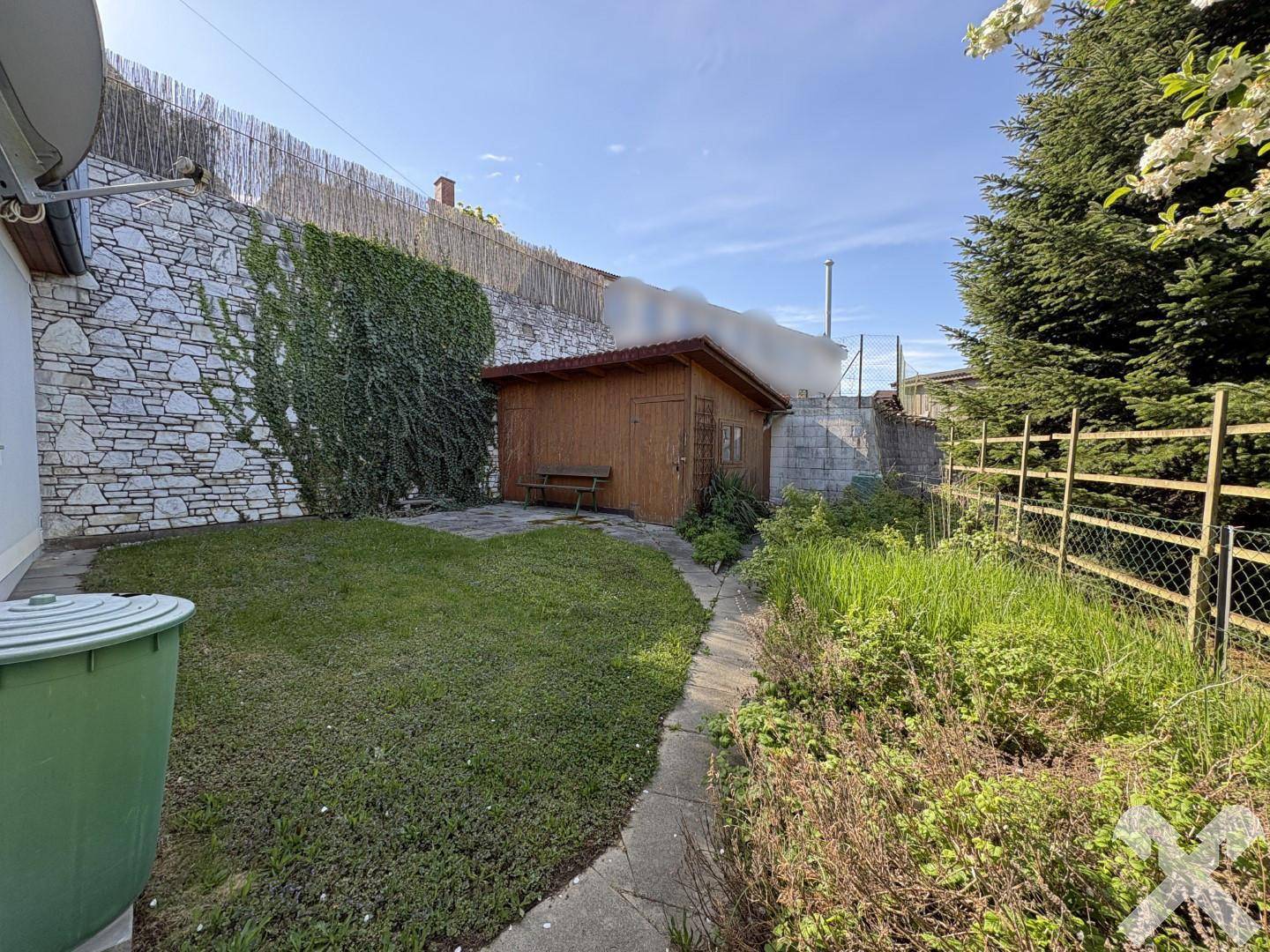 Hinterer Gartenbereich mit Rasenfläche, Gartenhaus und Steinmauer, ideal für Erholung im Freien.