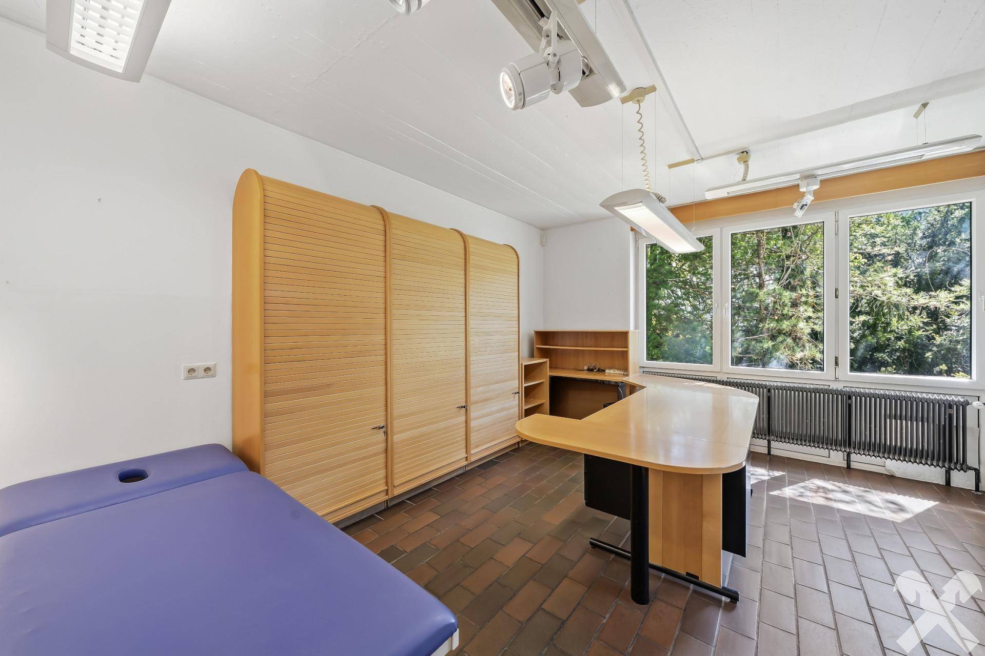 Helles Arbeitszimmer mit großen Fenstern, Holzboden und einer Behandlungsliege.