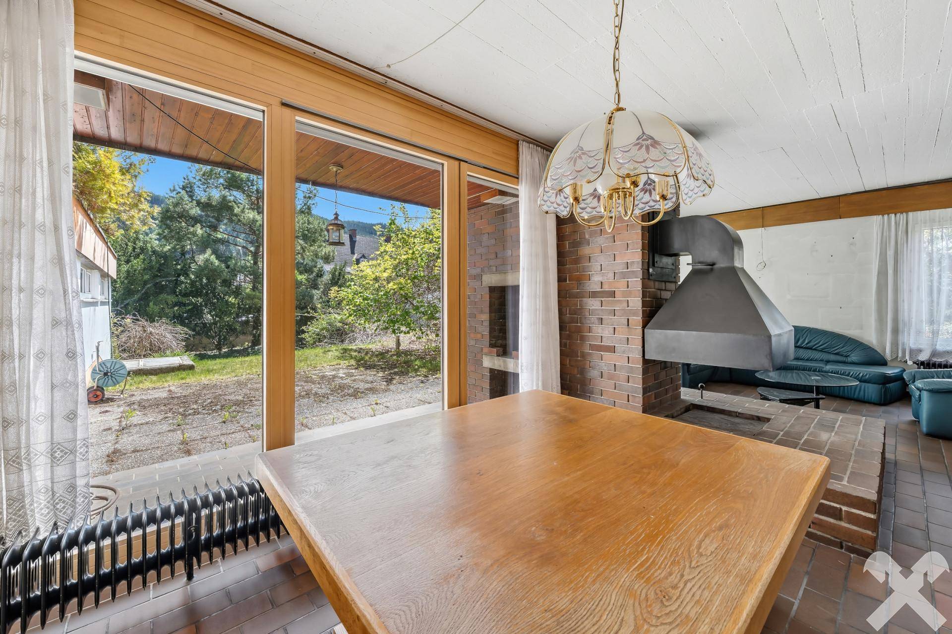 Essbereich mit Holztisch, Kamin und großen Fenstern mit Blick ins Grüne.