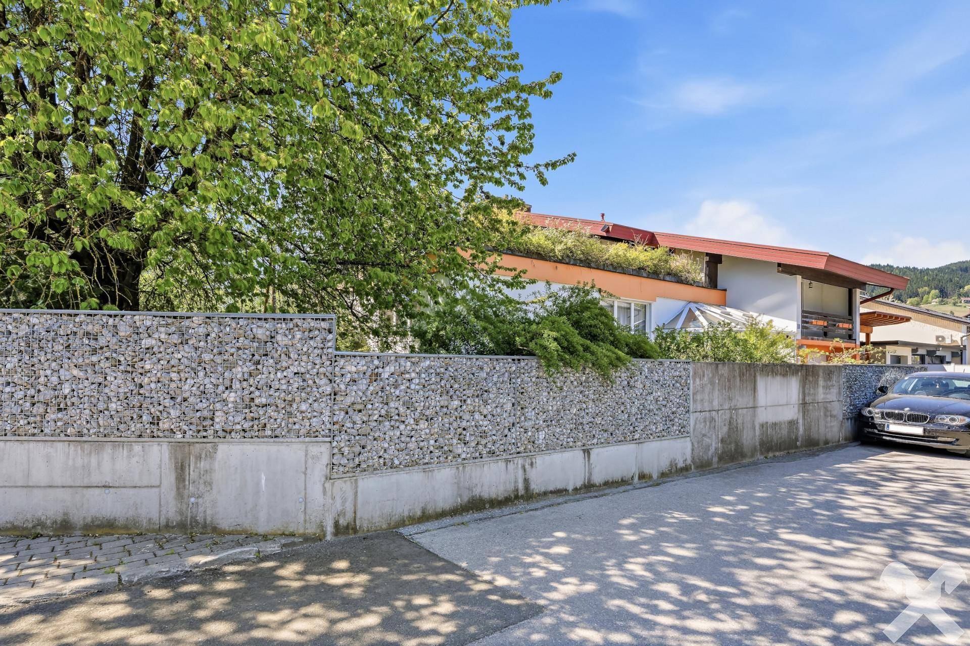 Blick von der Straße auf das Wohnhaus mit Gabionenmauer und geparktem Fahrzeug.