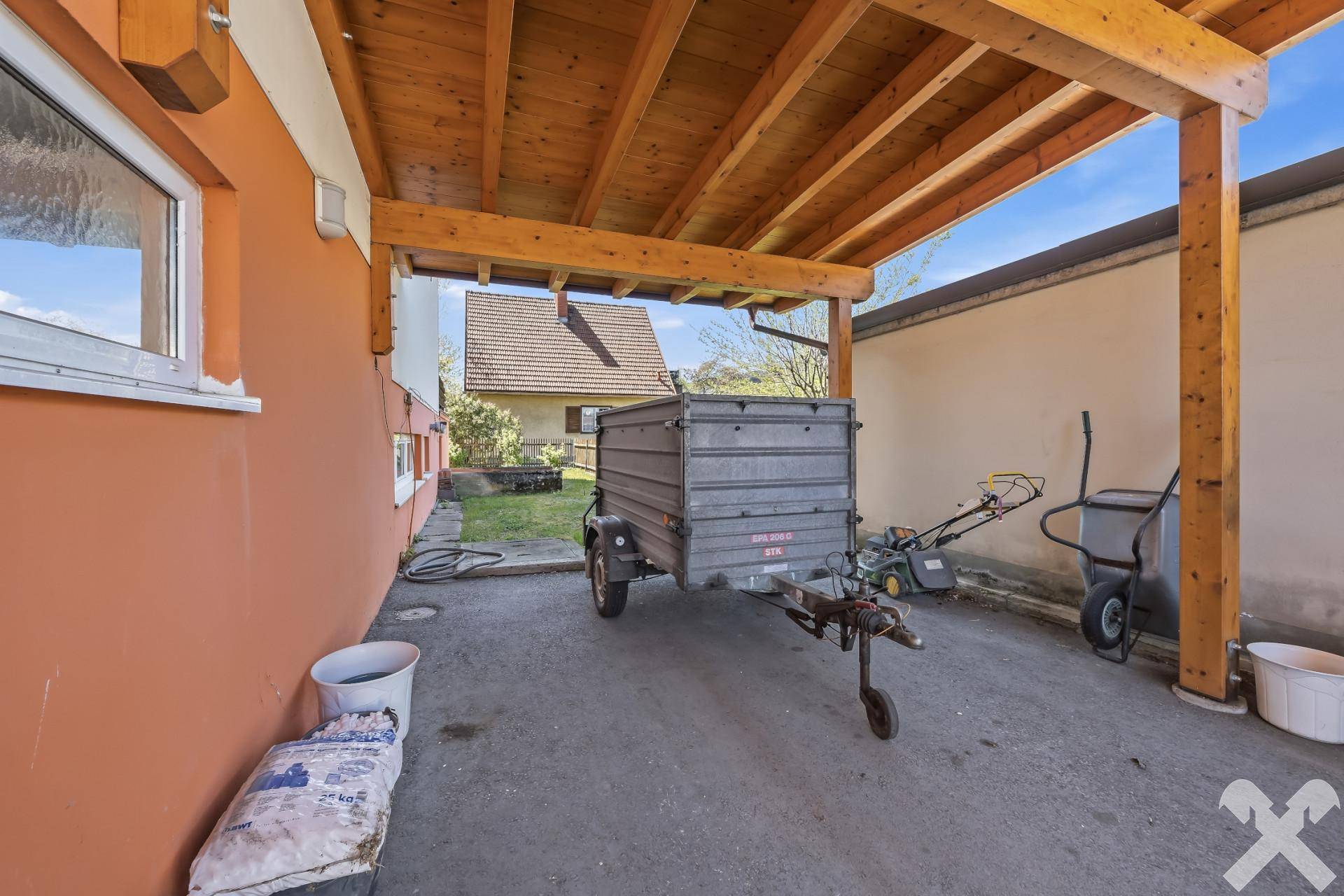 Überdachter Carport mit Holzbalkenkonstruktion, genutzt als Abstellplatz für Anhänger.