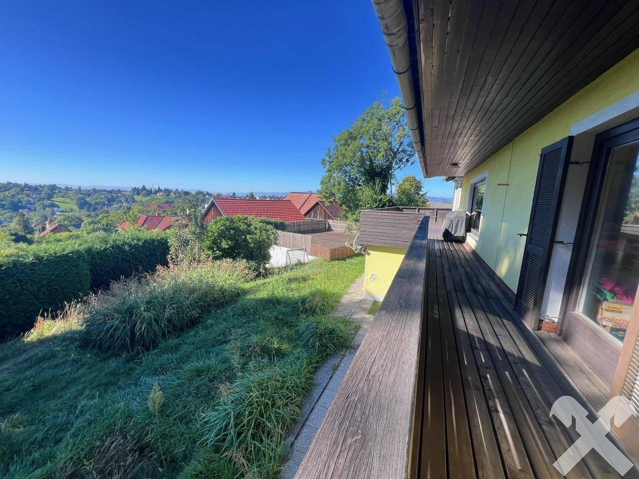 Langer Holzbalkon entlang der Hausfassade mit Blick auf die grüne Umgebung und entfernte Hügel.