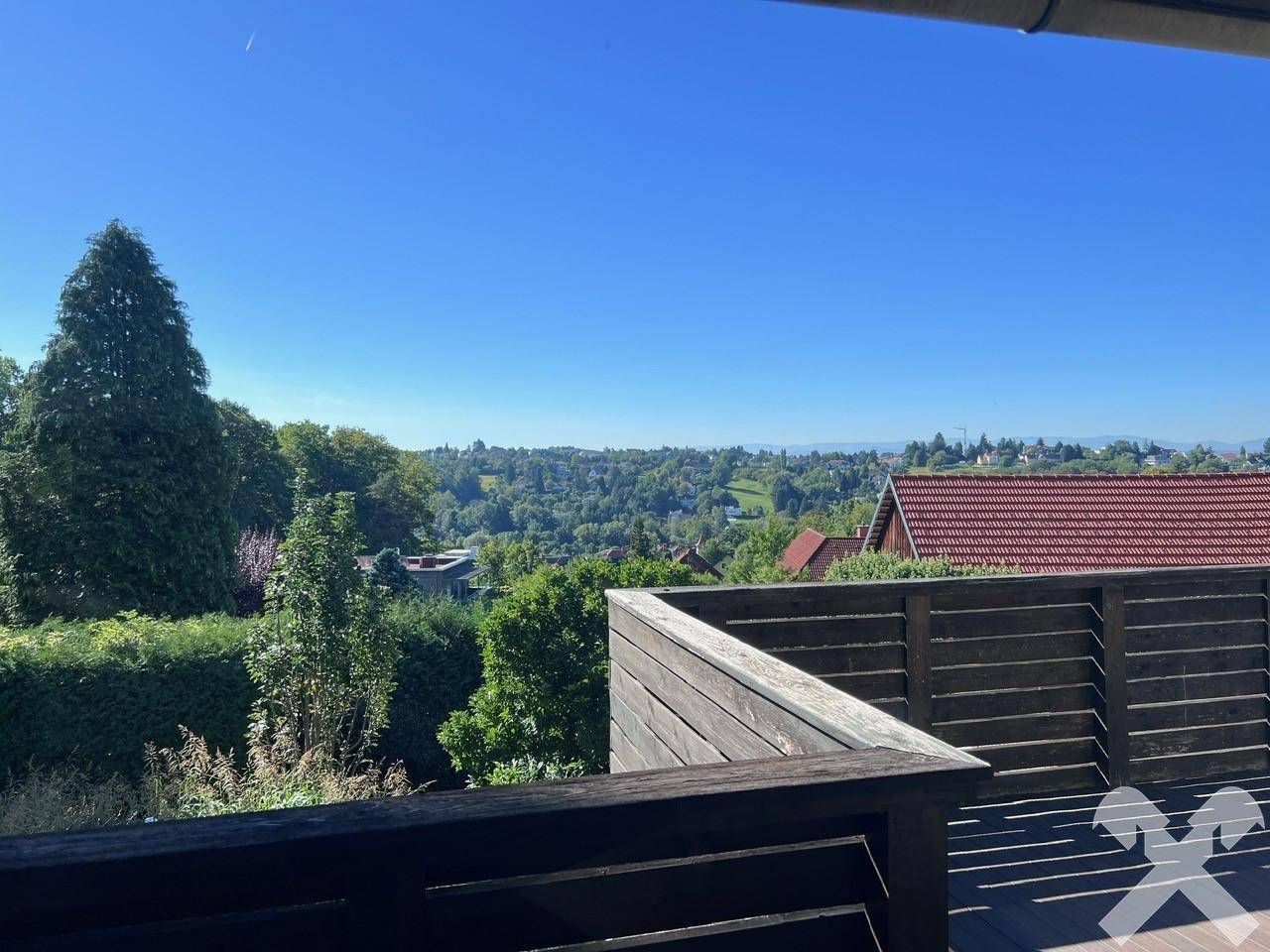 Großer Holzbalkon mit weitem Blick über die grüne Landschaft und Dächer unter klarem Himmel.