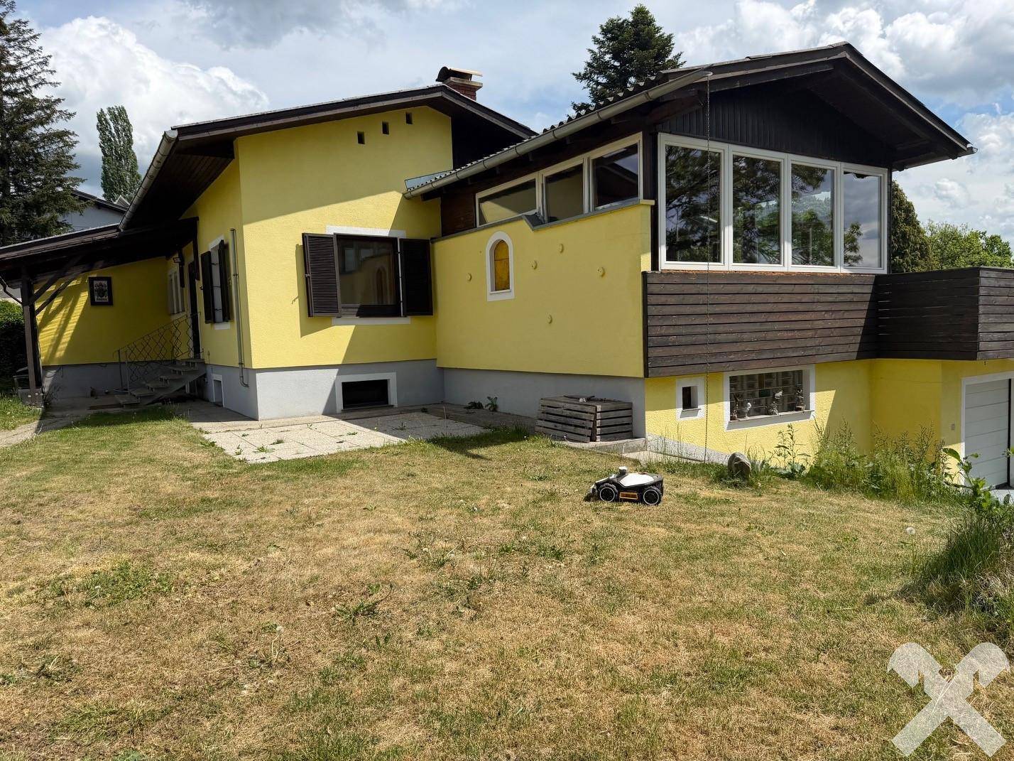 Seitenansicht des gelben Hauses mit großem Rasen und einem Mähroboter im Gartenbereich.