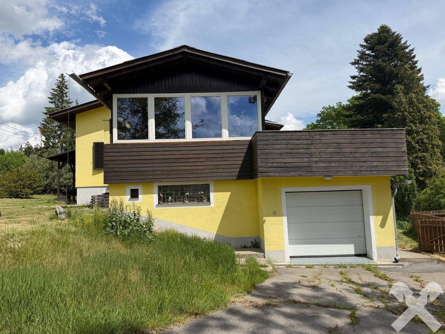 Gelbes Haus mit Garage und Balkon, umgeben von grüner Wiese unter blauem Himmel.